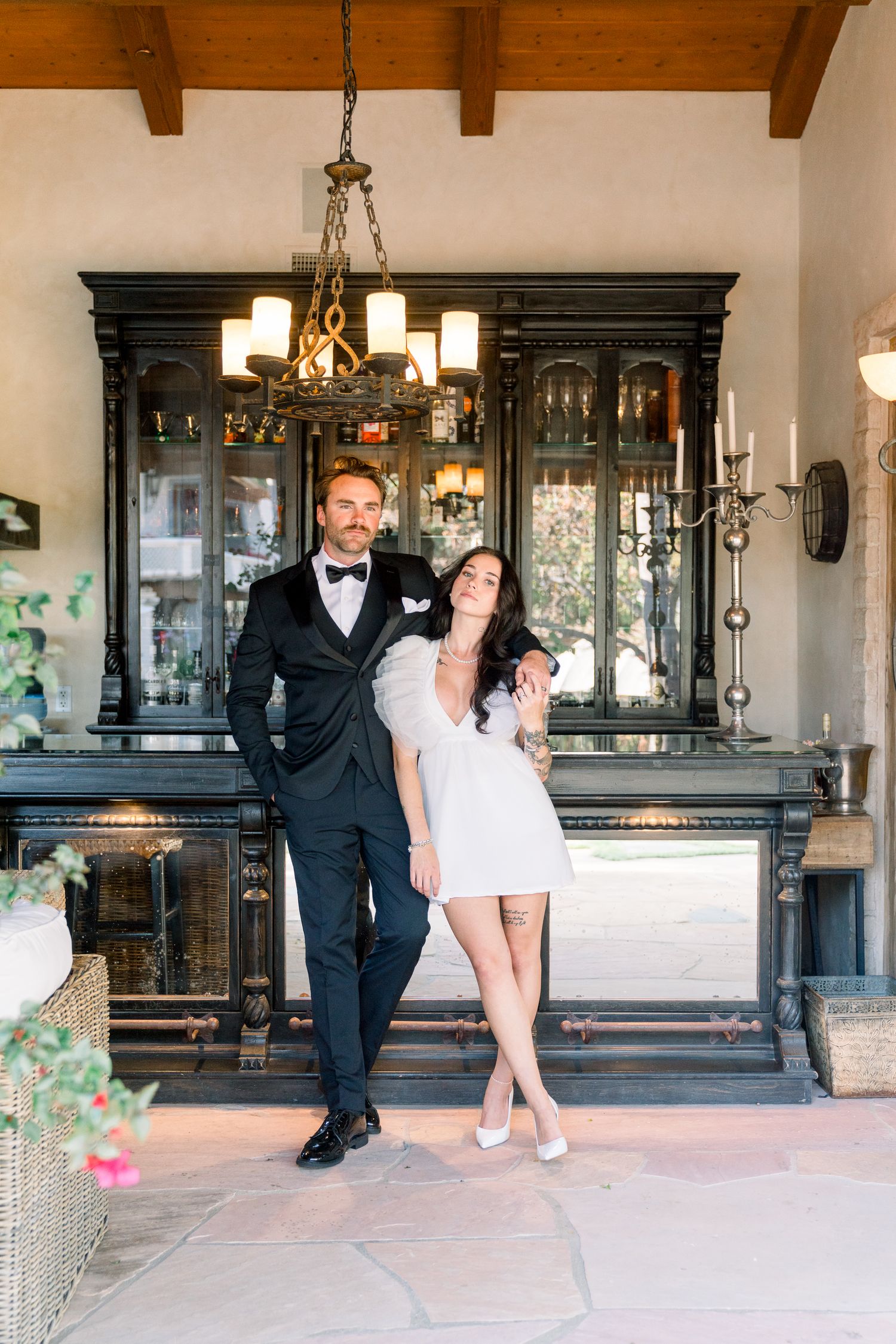 A couple poses together in formal attire against an elegant dark wood cabinet with a chandelier overhead.