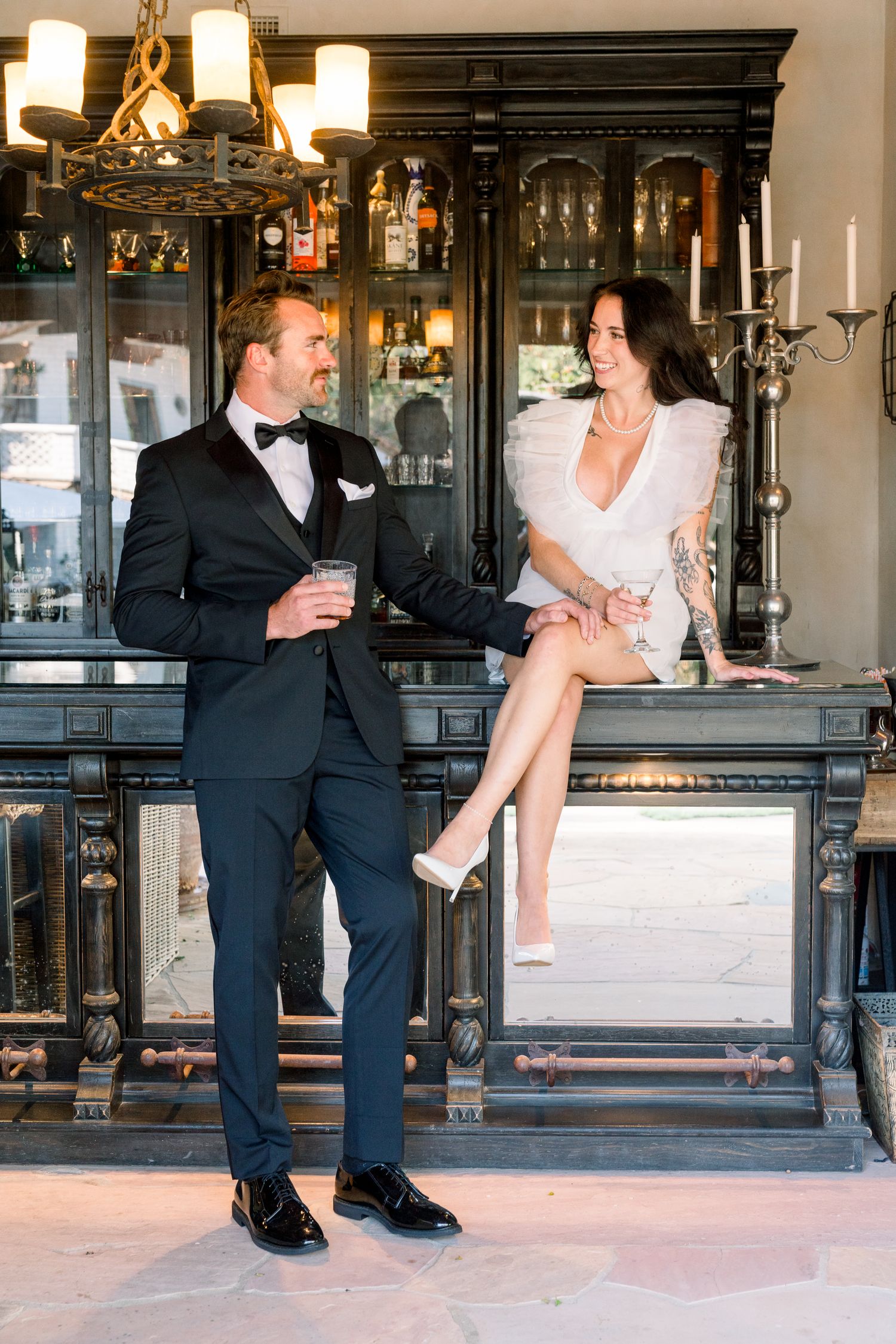 Elegant couple in formal attire sharing drinks at vintage bar with chandelier and dark wood details.