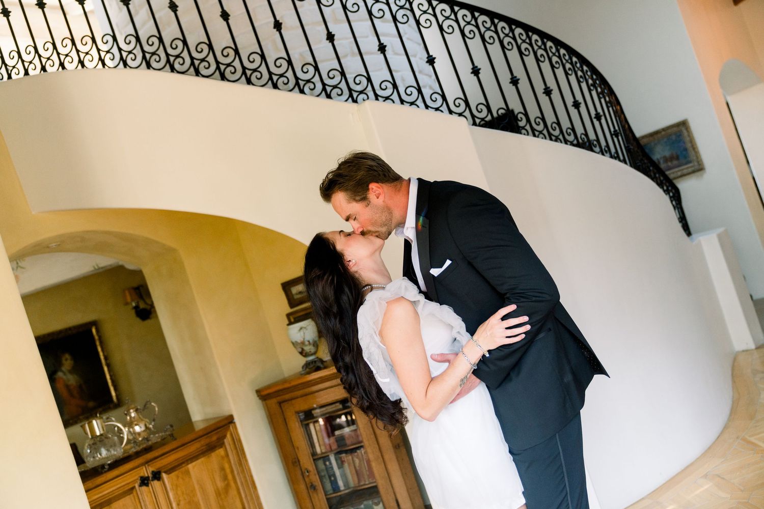 A bride and groom share a romantic kiss by an elegant curved staircase with wrought iron railings.