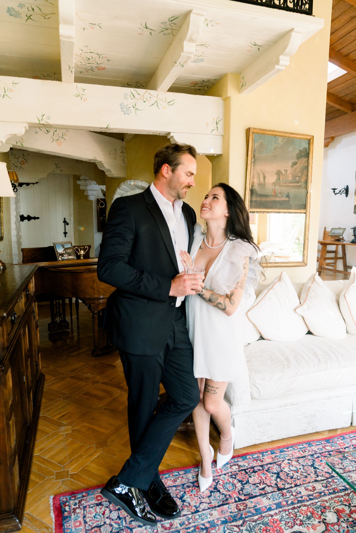 A couple shares an intimate moment in a rustic interior with white walls, wooden beams, and a decorative rug.