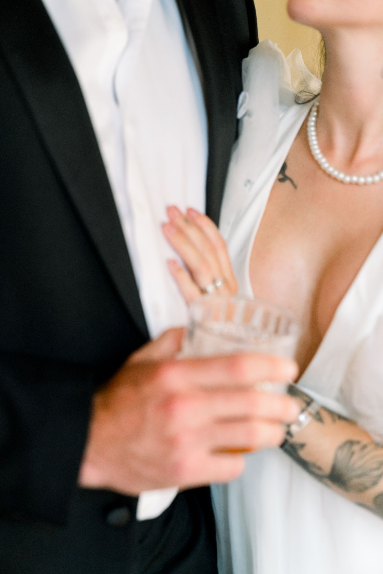 Close-up of couple in formal wear with pearls and tattoos holding glass in soft romantic lighting.