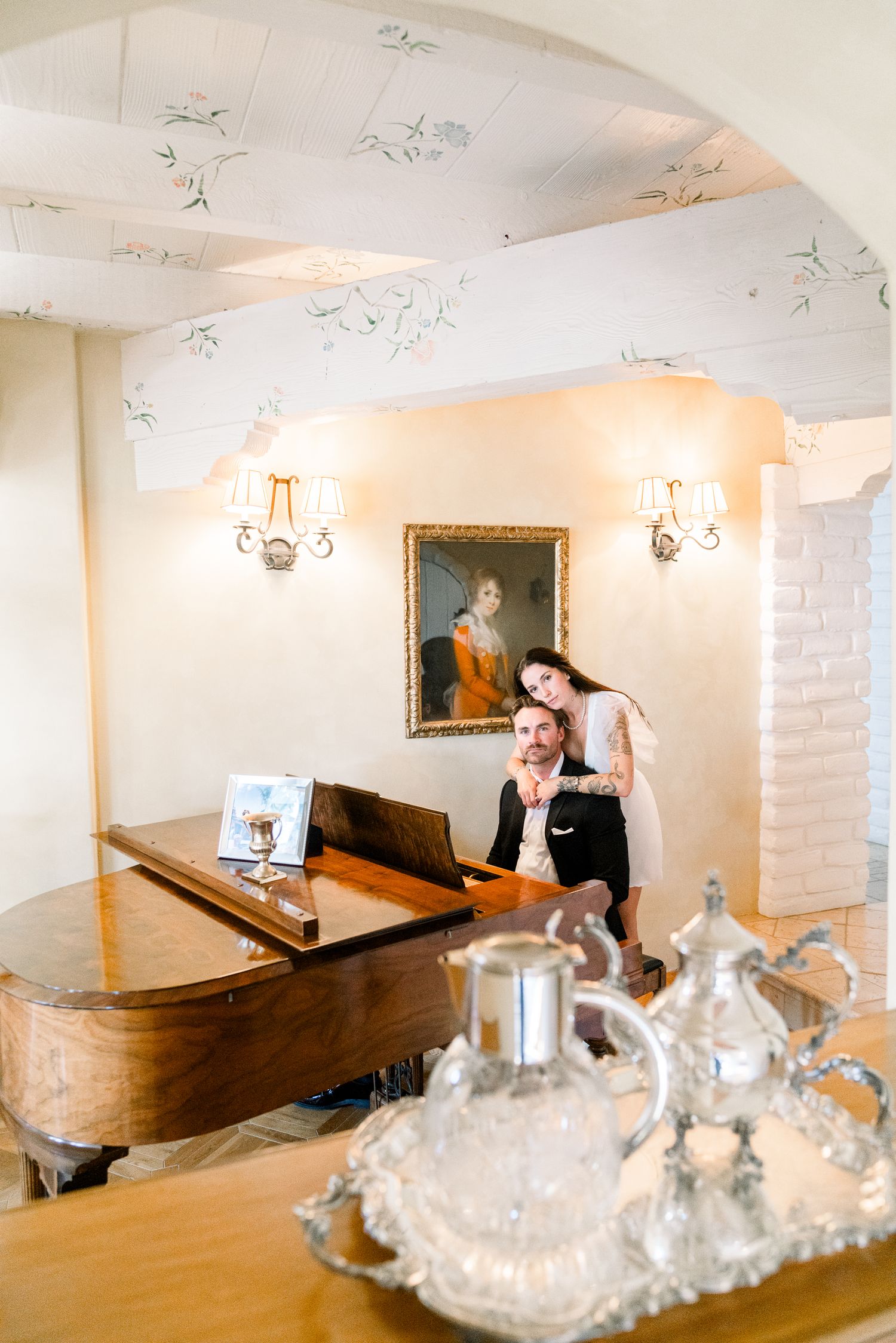 A grand room with a wooden piano, intricate ceiling details, and a silver tea service set on display.