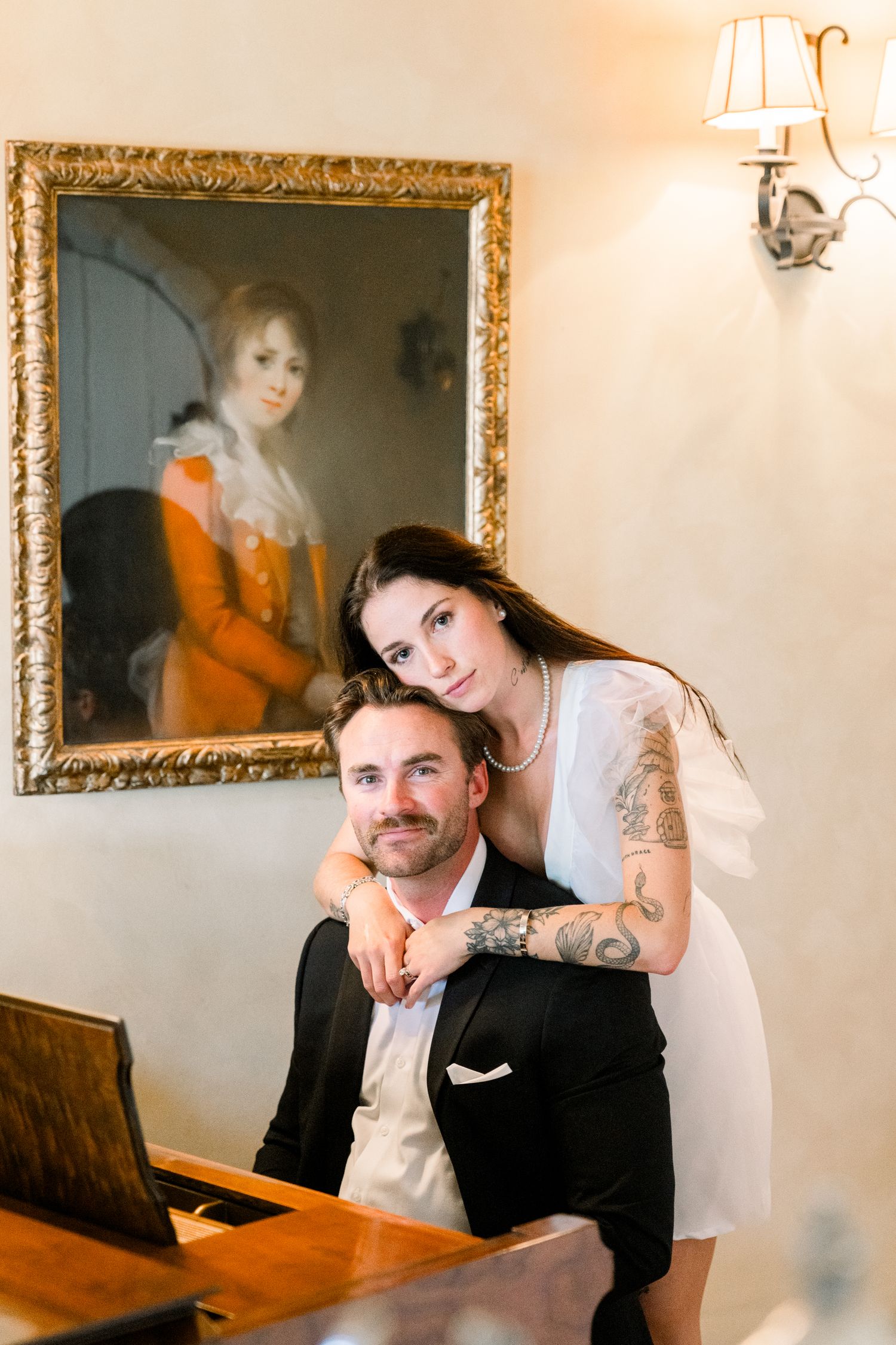 A couple poses together at an ornate wooden piano beneath a classical portrait in an elegant interior setting.
