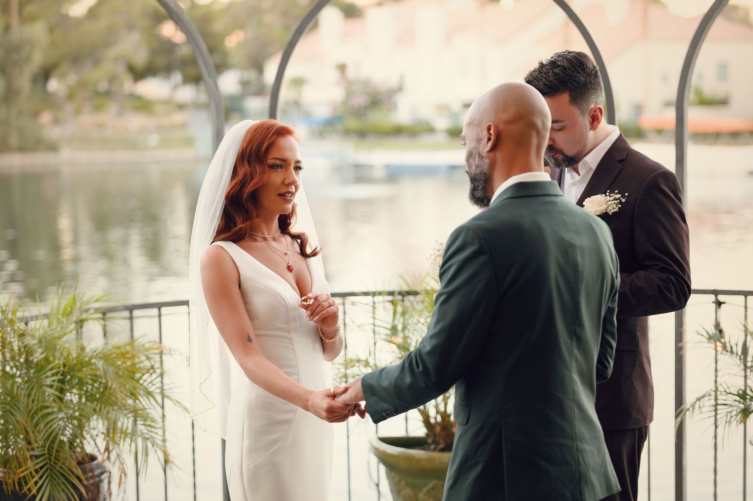 A bride and groom hold hands during their wedding ceremony by a scenic lake, as the officiant reads vows in the background.