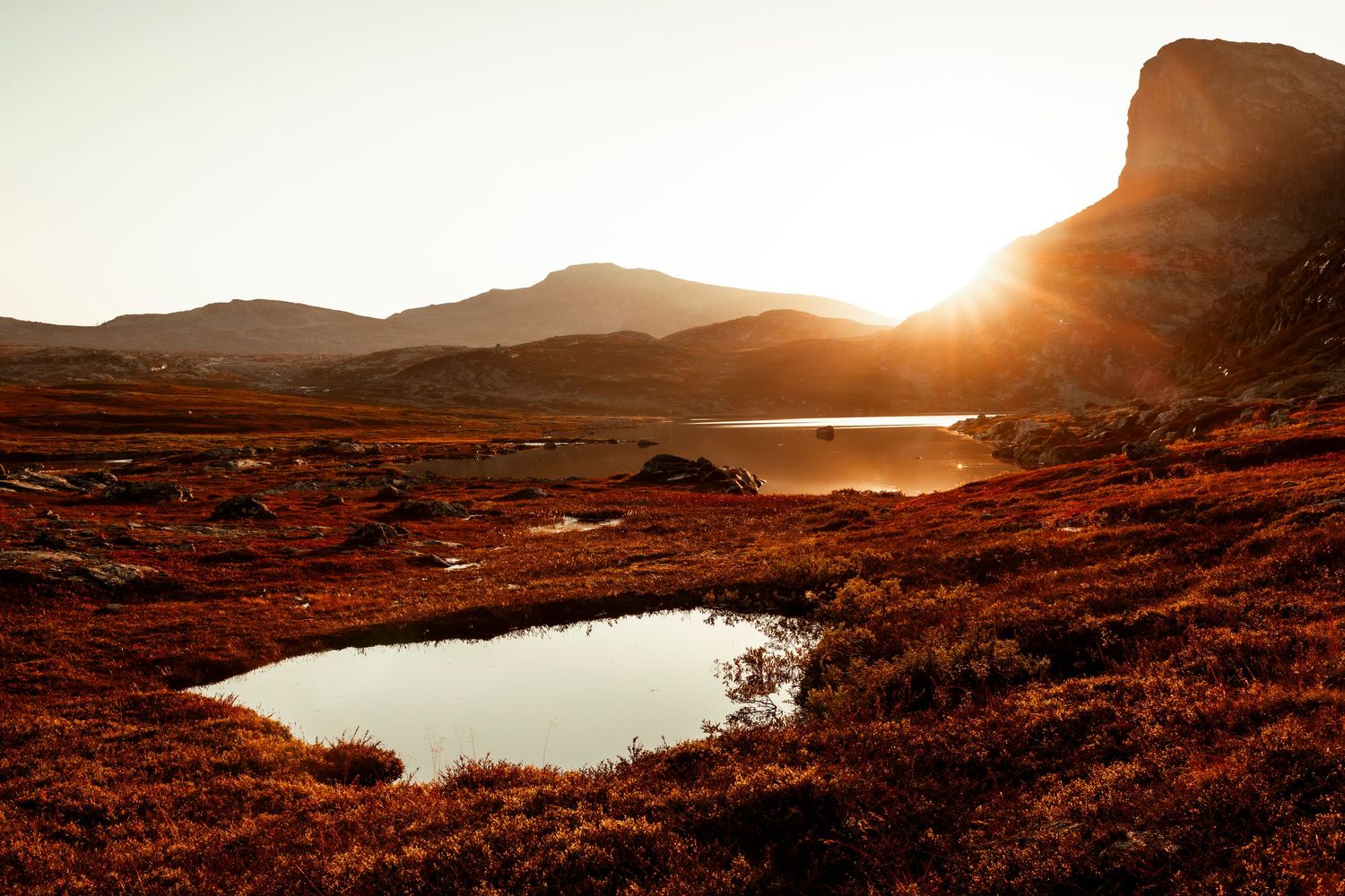 Dramatic sunset illuminates rustic mountain landscape with two small lakes reflecting golden light in reddish terrain.
