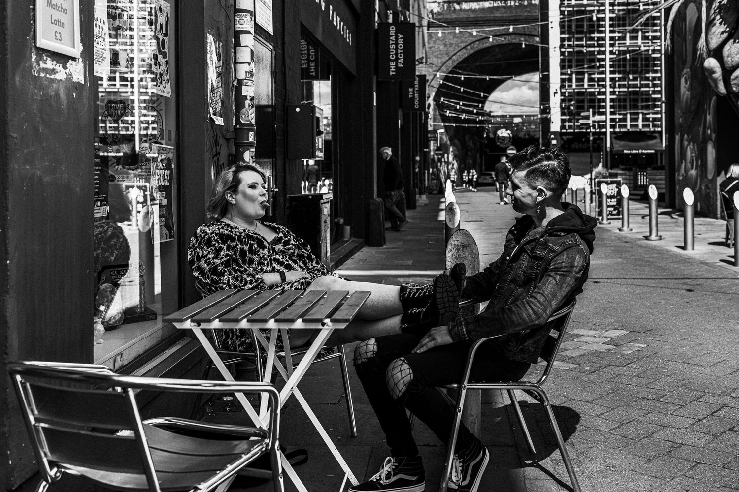 Two people share a table at an outdoor cafe on a city street with an urban backdrop in black and white.