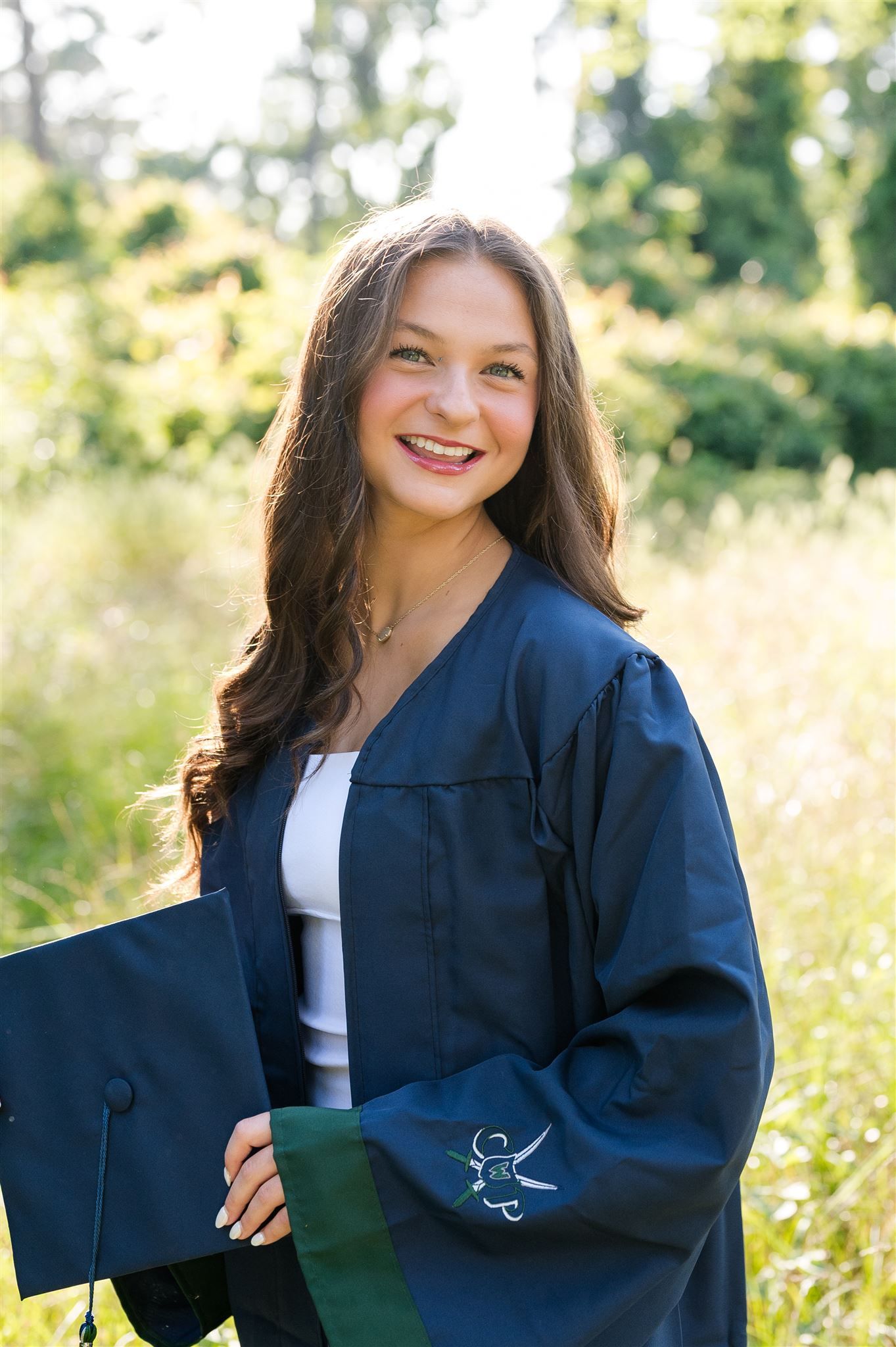 Graduate wearing navy blue academic gown smiles while posing in a sunny meadow with tall grass in background.