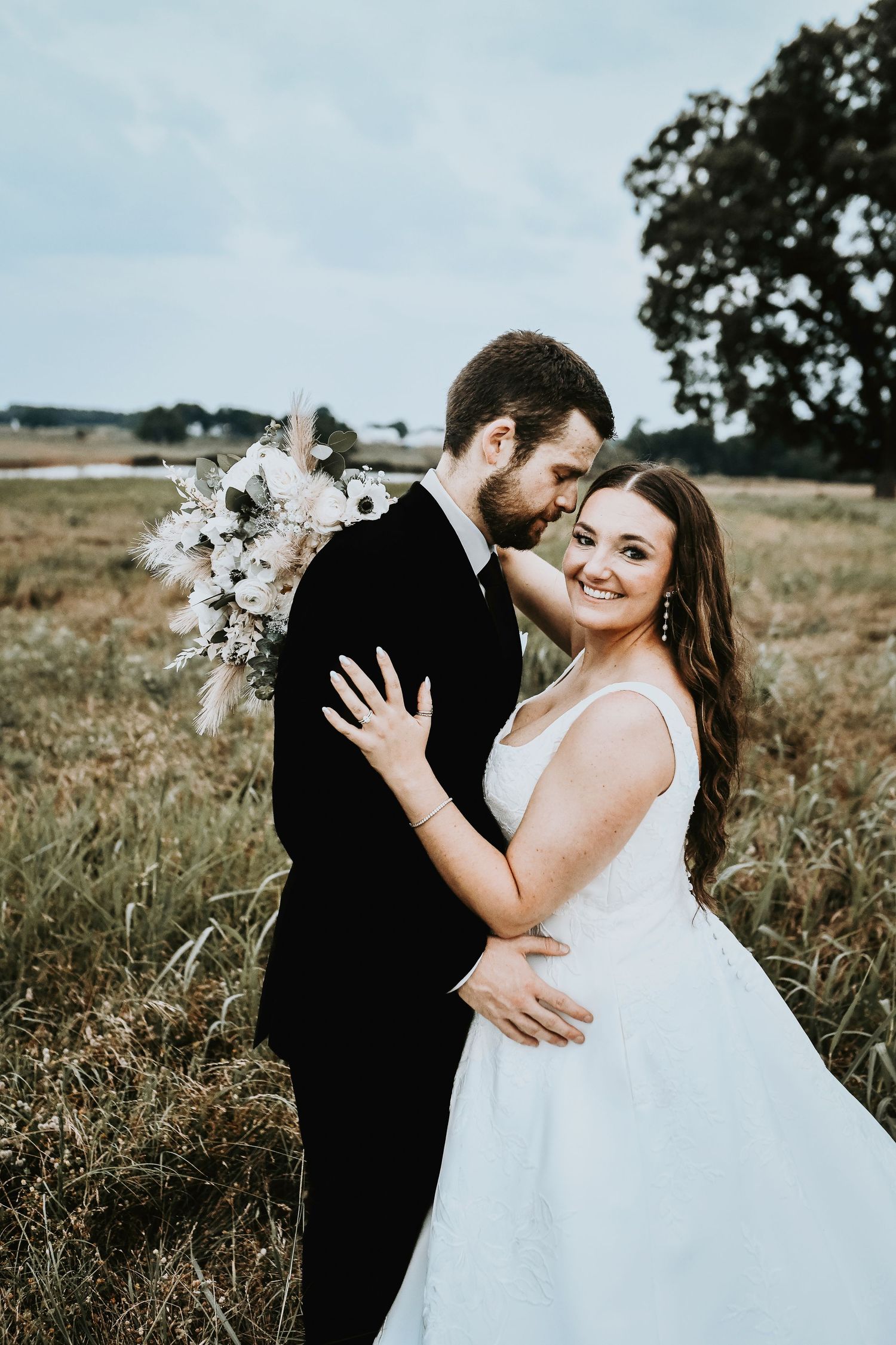 A bride and groom romantically embrace in a field of spring grass as the bride smiles at the camera for their wedding photos
