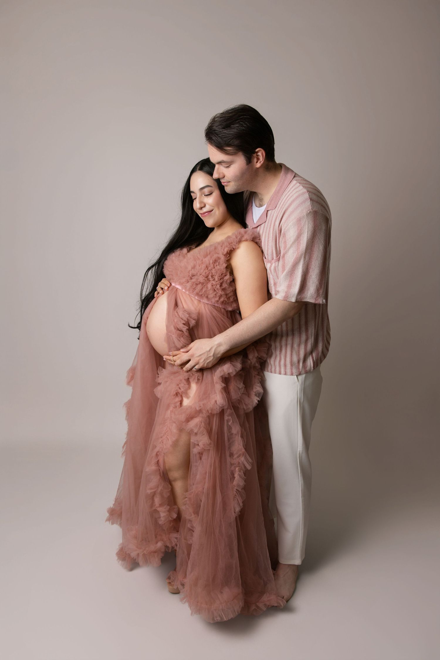 A couple in matching pink outfits embraces during a maternity photoshoot against a neutral backdrop.
