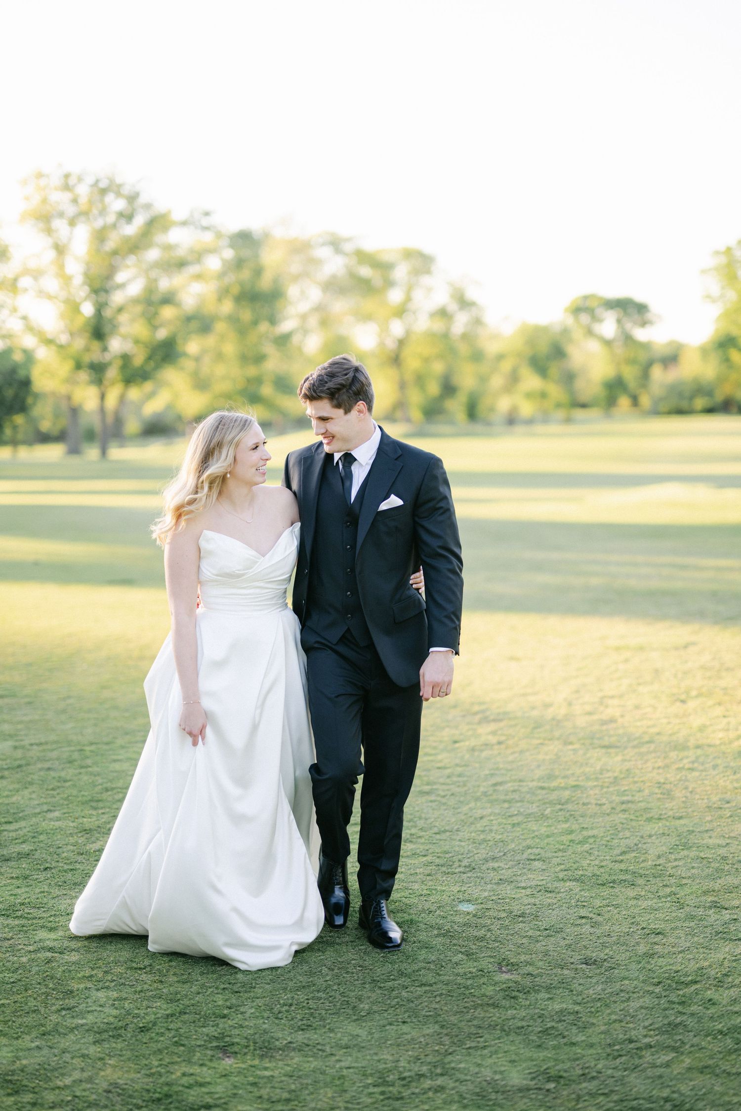 A couple walking together on a lush green golf course at golden hour, the bride in white and groom in black suit.
