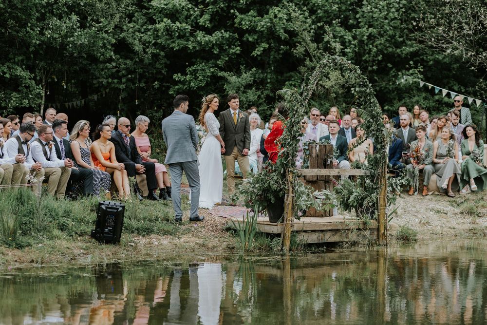 An outdoor wedding ceremony takes place beside a tranquil pond with guests seated on wooden benches in a natural setting.