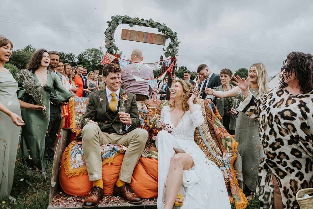 A wedding celebration unfolds with lively guests seated on orange pumpkin-themed chairs at an outdoor fall ceremony.