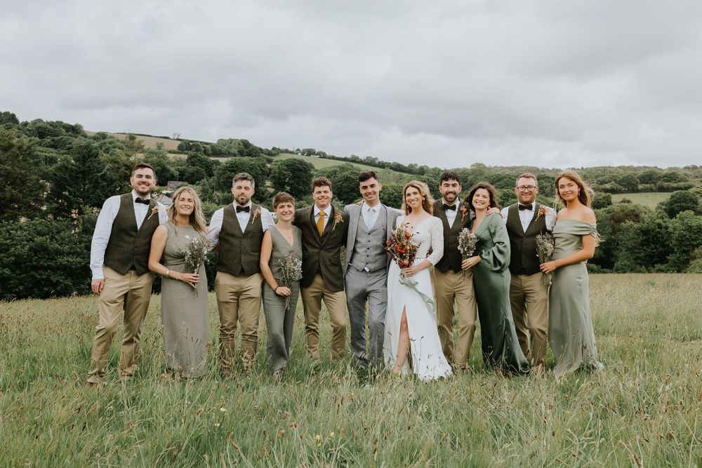 Wedding party poses together in an outdoor field with rolling hills and cloudy skies in the background.