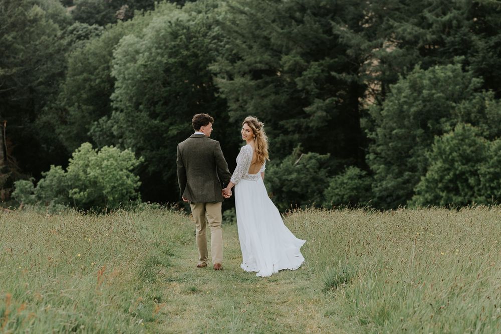 Bride and groom dance playfully in a scenic meadow surrounded by lush green trees on their wedding day.