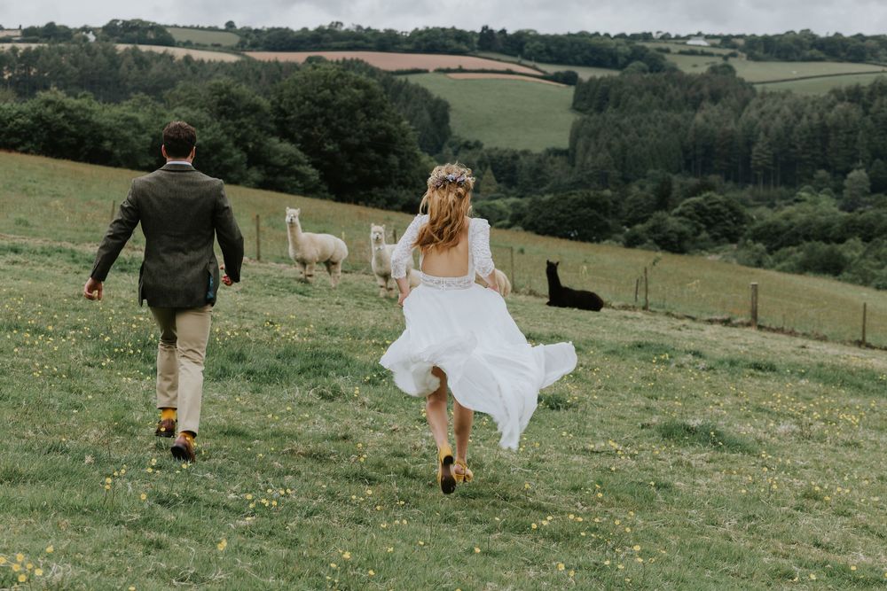 Two figures walk across a green hillside meadow with a flowing white dress catching the wind against a pastoral landscape.