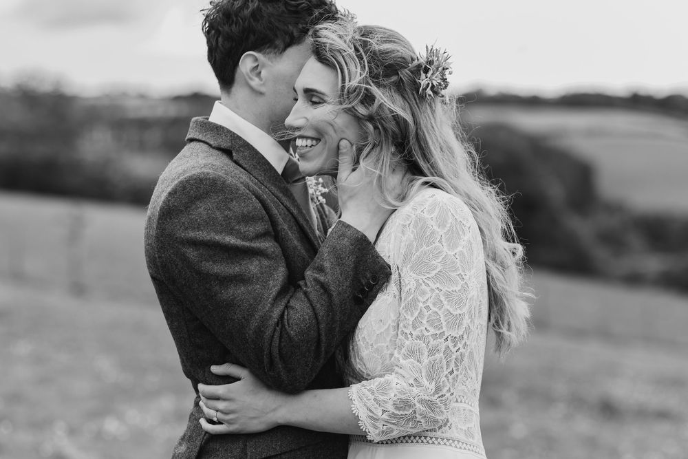 Black and white photo of couple embracing outdoors, bride in lace dress and groom in tweed jacket sharing intimate moment.