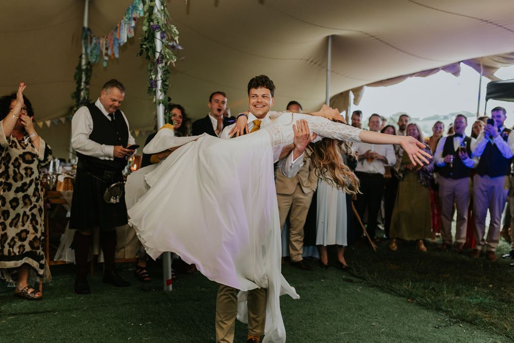 A couple dances their first dance at a wedding reception under a tent while guests watch and take photos.