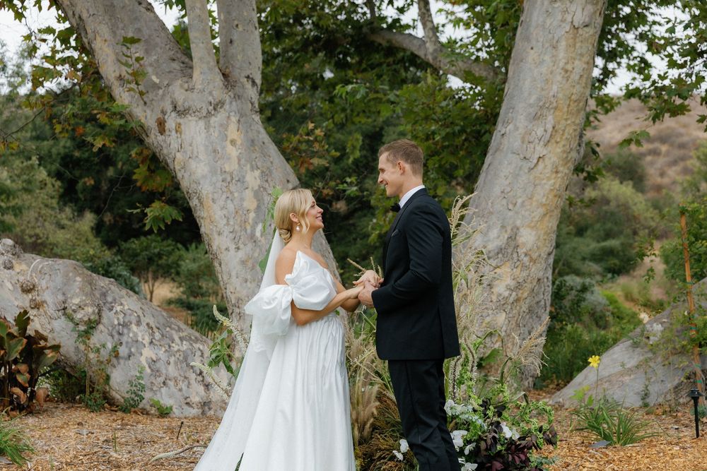 A couple stands together under large sycamore trees during their outdoor wedding ceremony.