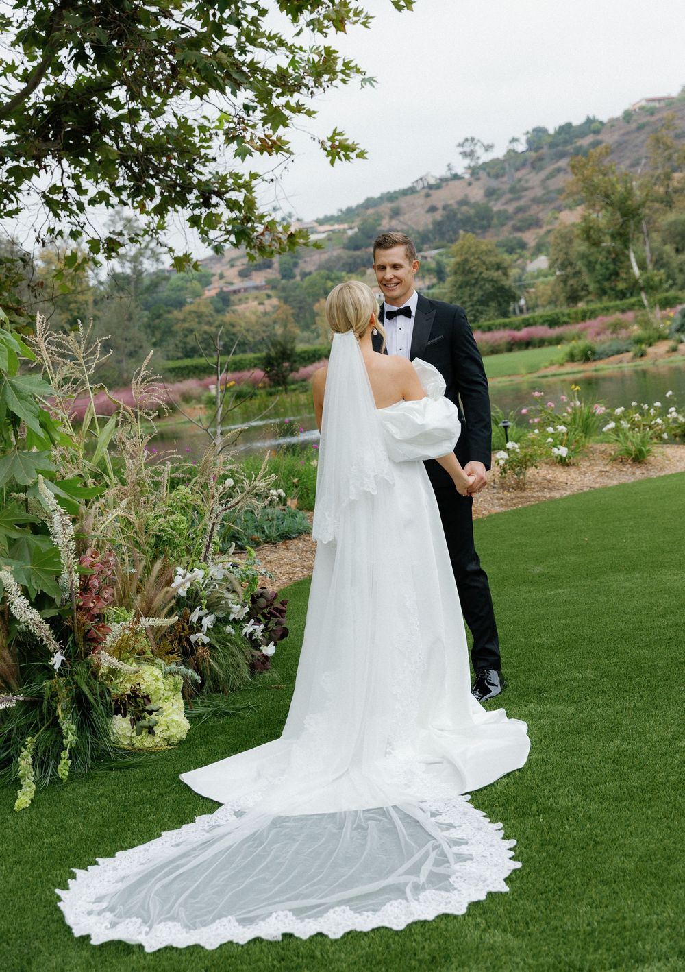 A wedding couple embraces on a manicured lawn with hills and gardens in the background, the bride's lace veil trailing behind.
