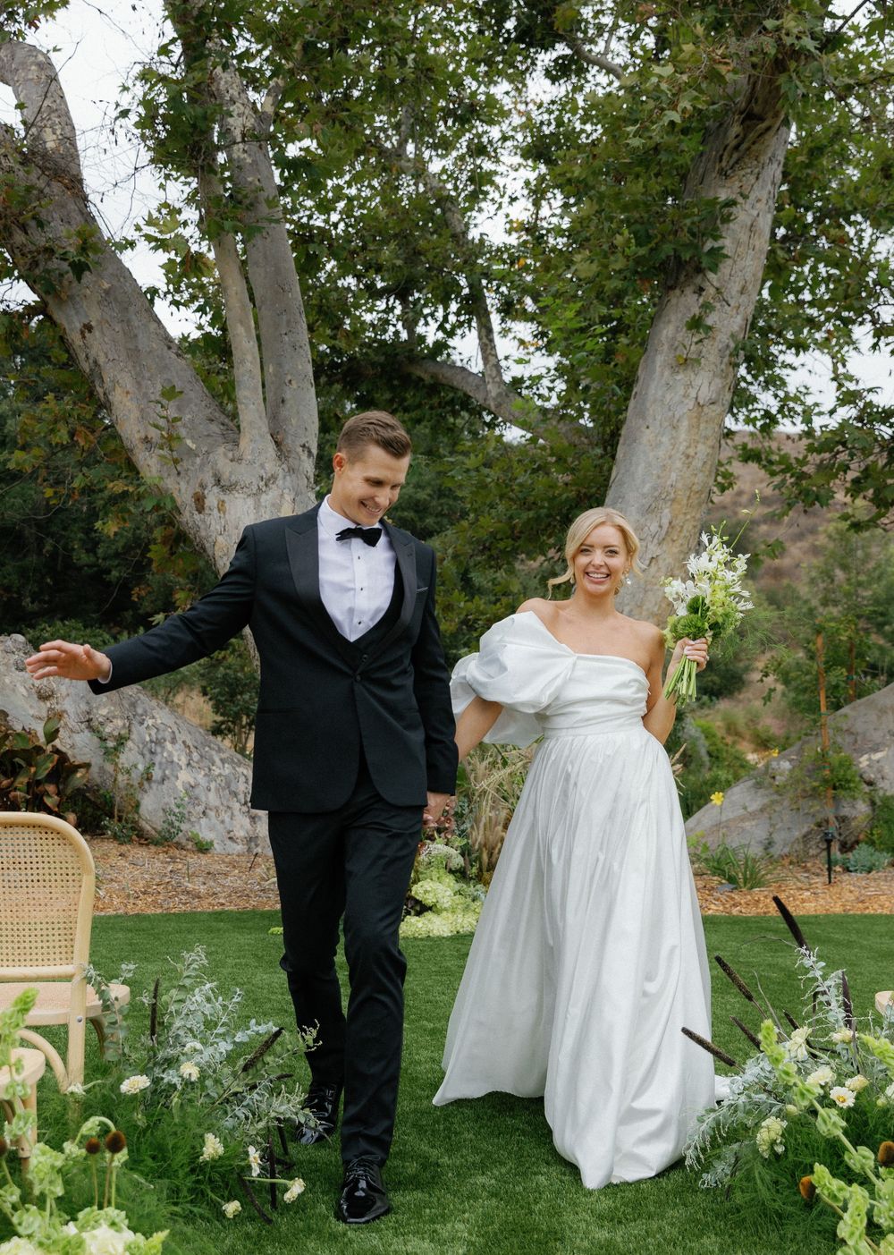 A couple in wedding attire stand together on a grass lawn with floral arrangements and a sycamore tree backdrop.