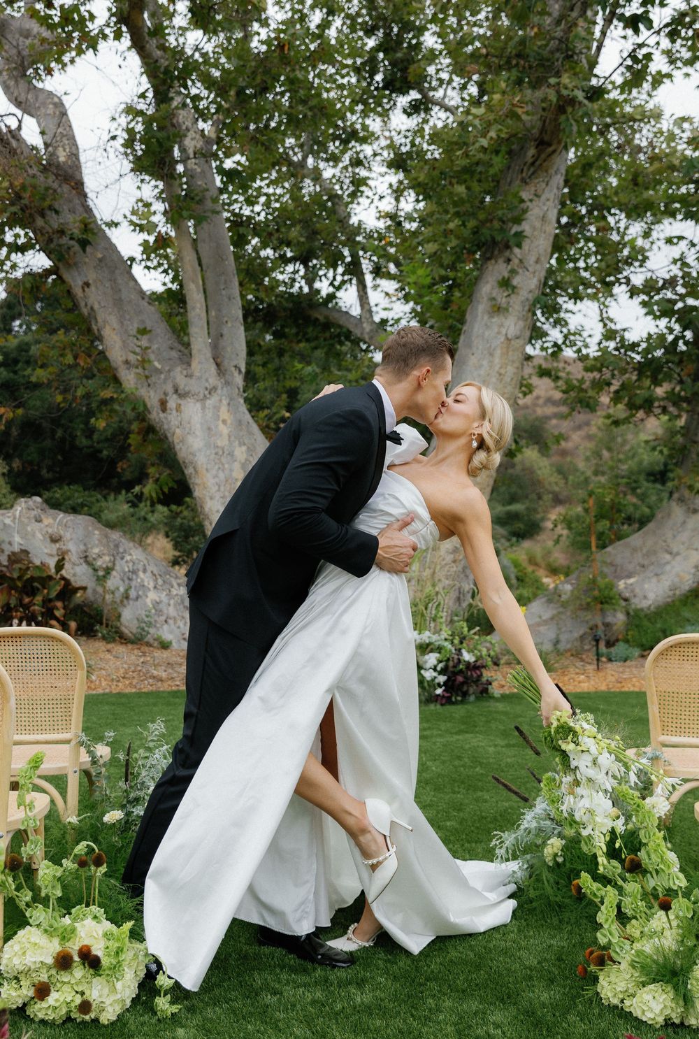 Romantic wedding couple shares a kiss under a majestic sycamore tree with wicker chairs and floral arrangements.