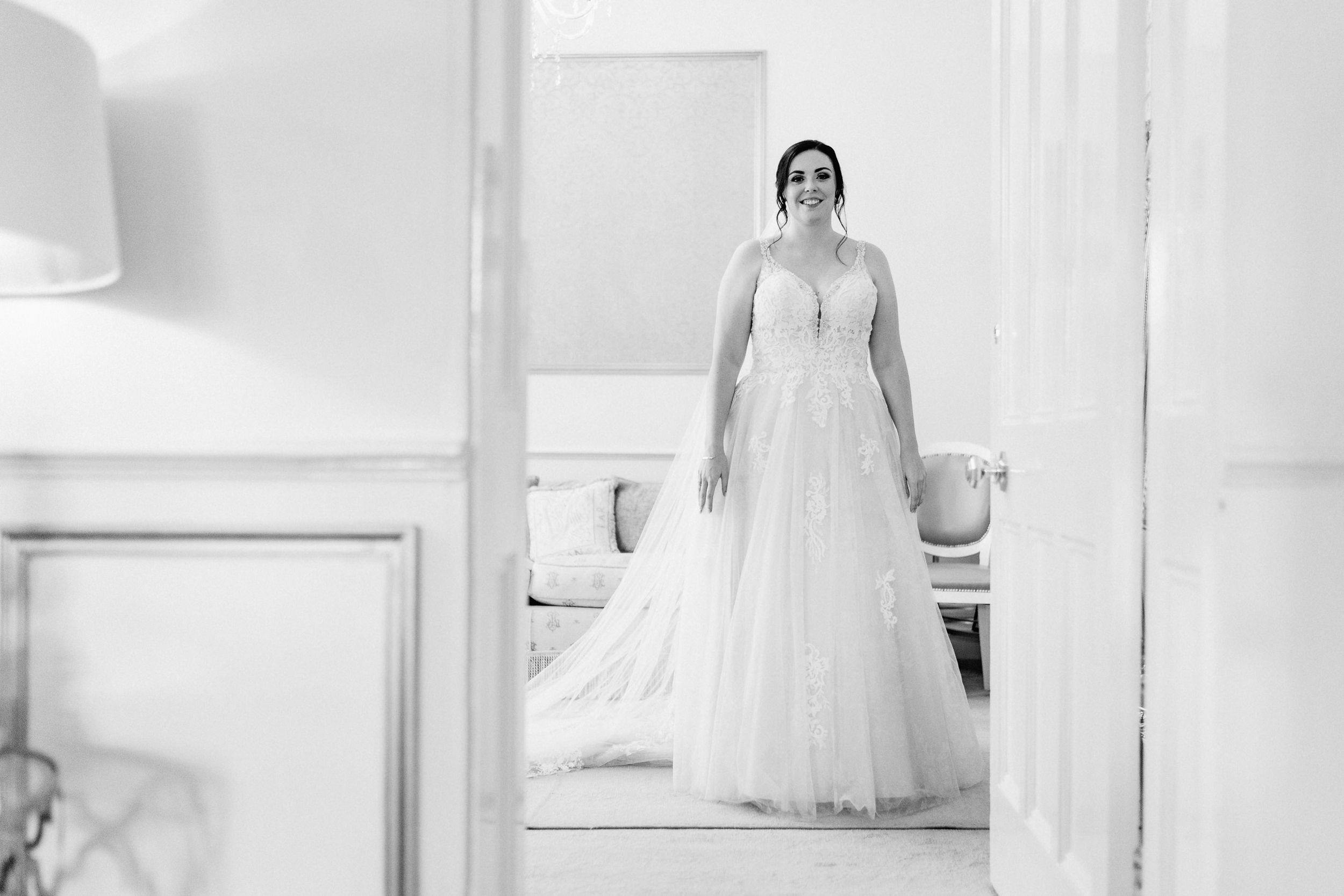 Black and white reflection of a bride in a flowing white wedding gown and veil standing in an elegant room.