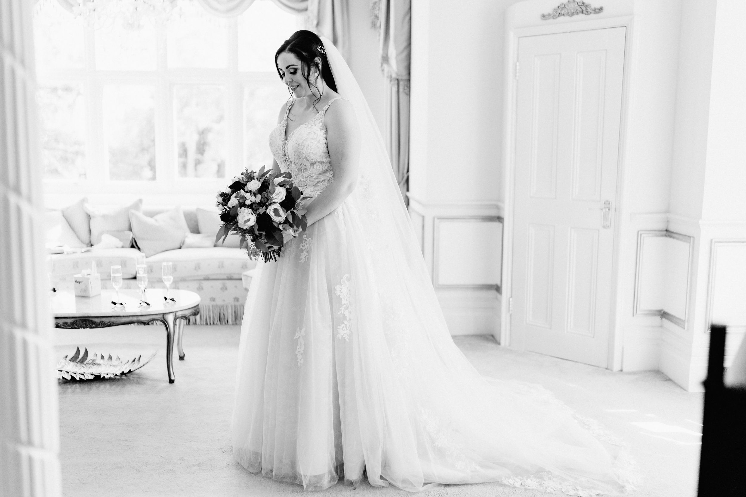 A black and white photo of a bride at Holmewood Hall in a flowing tulle wedding gown holding a lush floral bouquet.