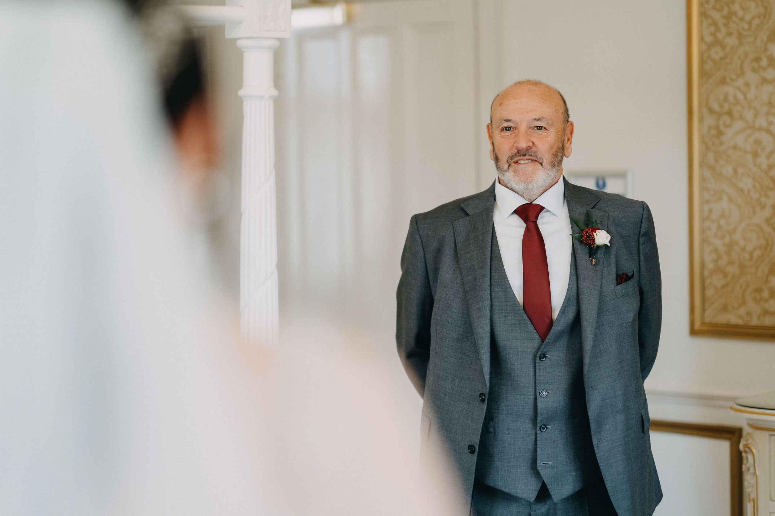 Wedding guest in a charcoal grey three-piece suit with red tie and boutonniere smiles in elegant venue hallway.
