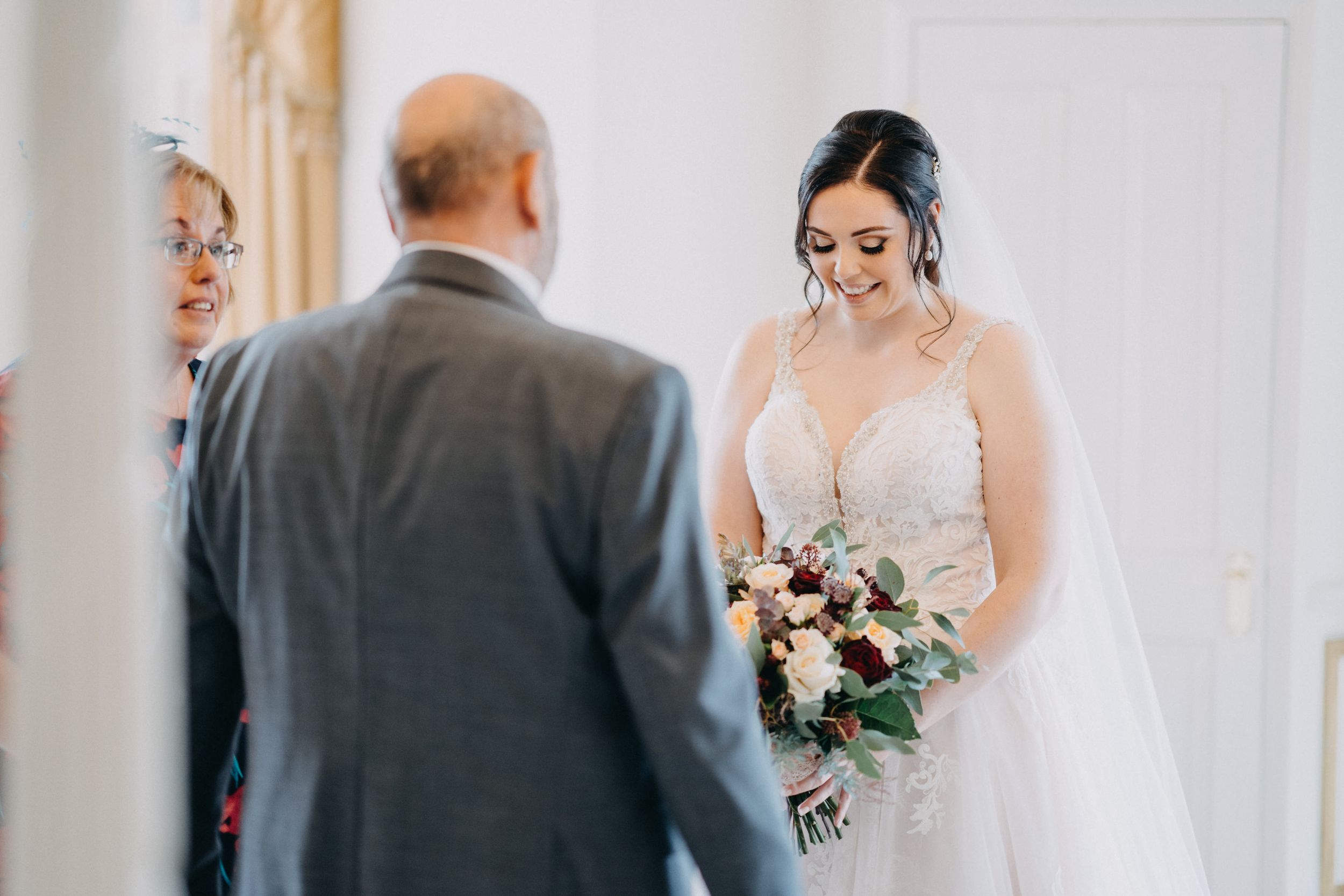 A smiling bride in a white lace wedding dress holds a burgundy and blush bouquet during her ceremony.