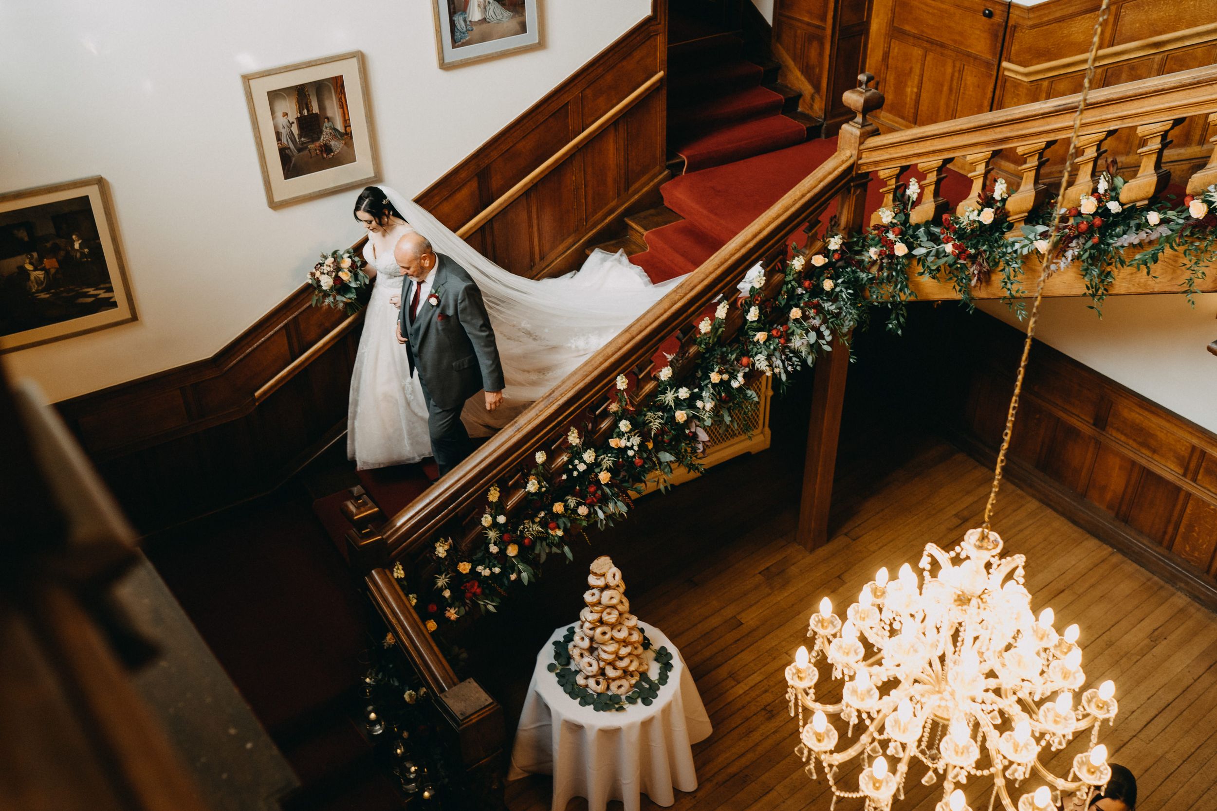Elegant Holmewood Hall with decorative garland on staircase, crystal chandelier, and wedding cake on round table.