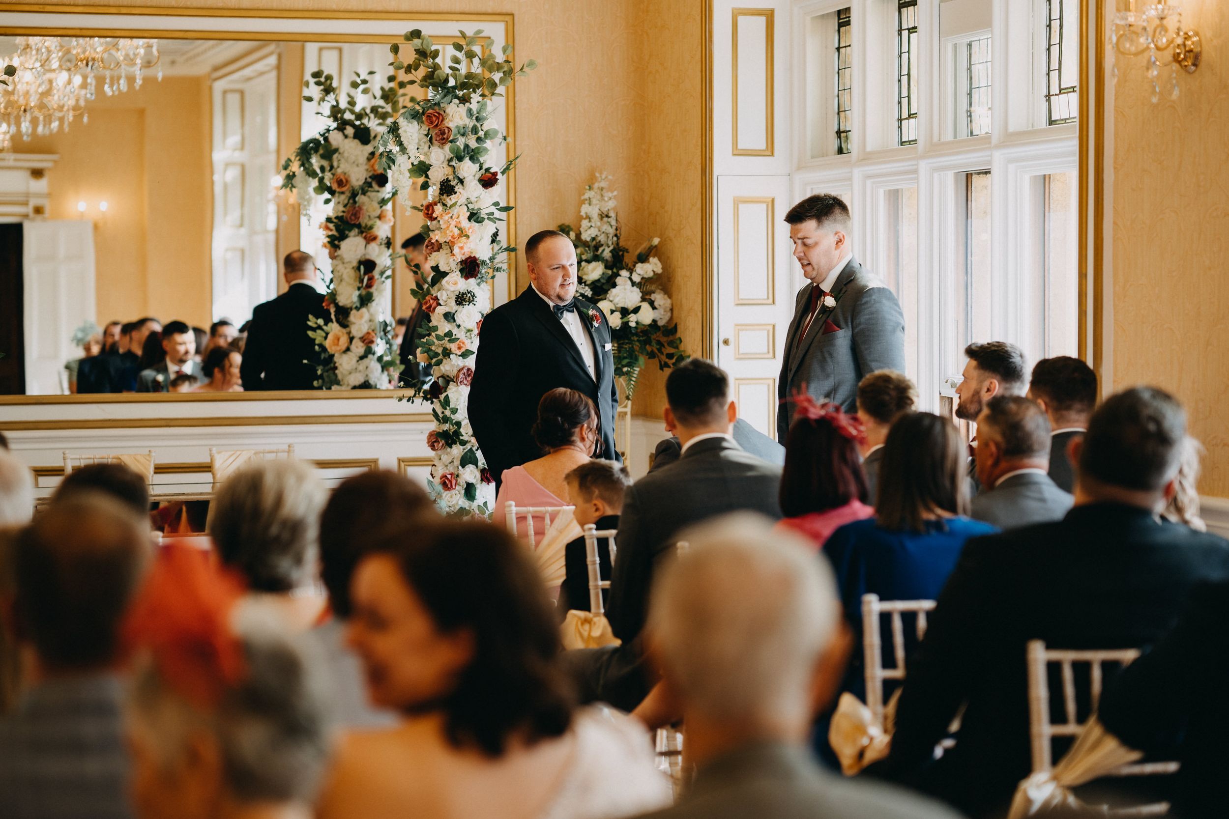 People gather for an elegant indoor ceremony at Holmewood Hall with floral decorations and white paneled walls in a classic venue setting.