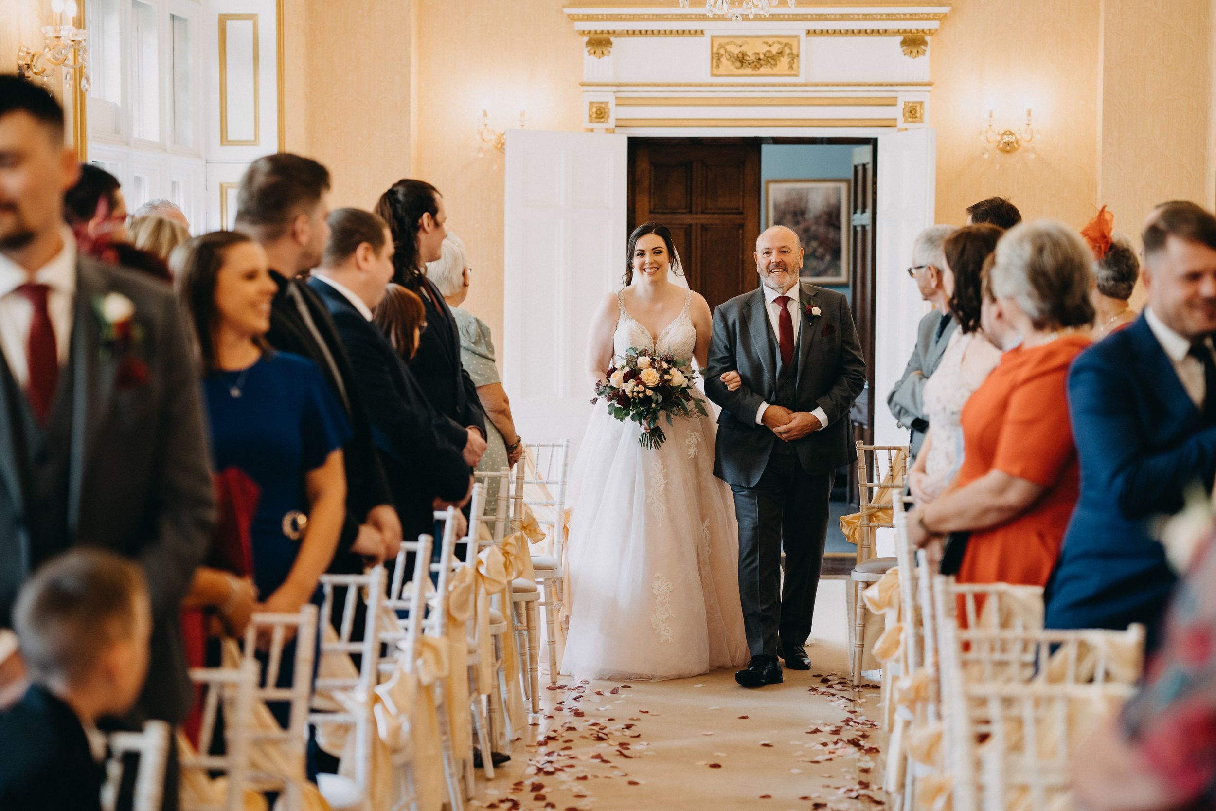 A wedding ceremony at Holmewood Hall, Peterborough in an elegant ballroom with white chairs and rose petals lining the aisle.