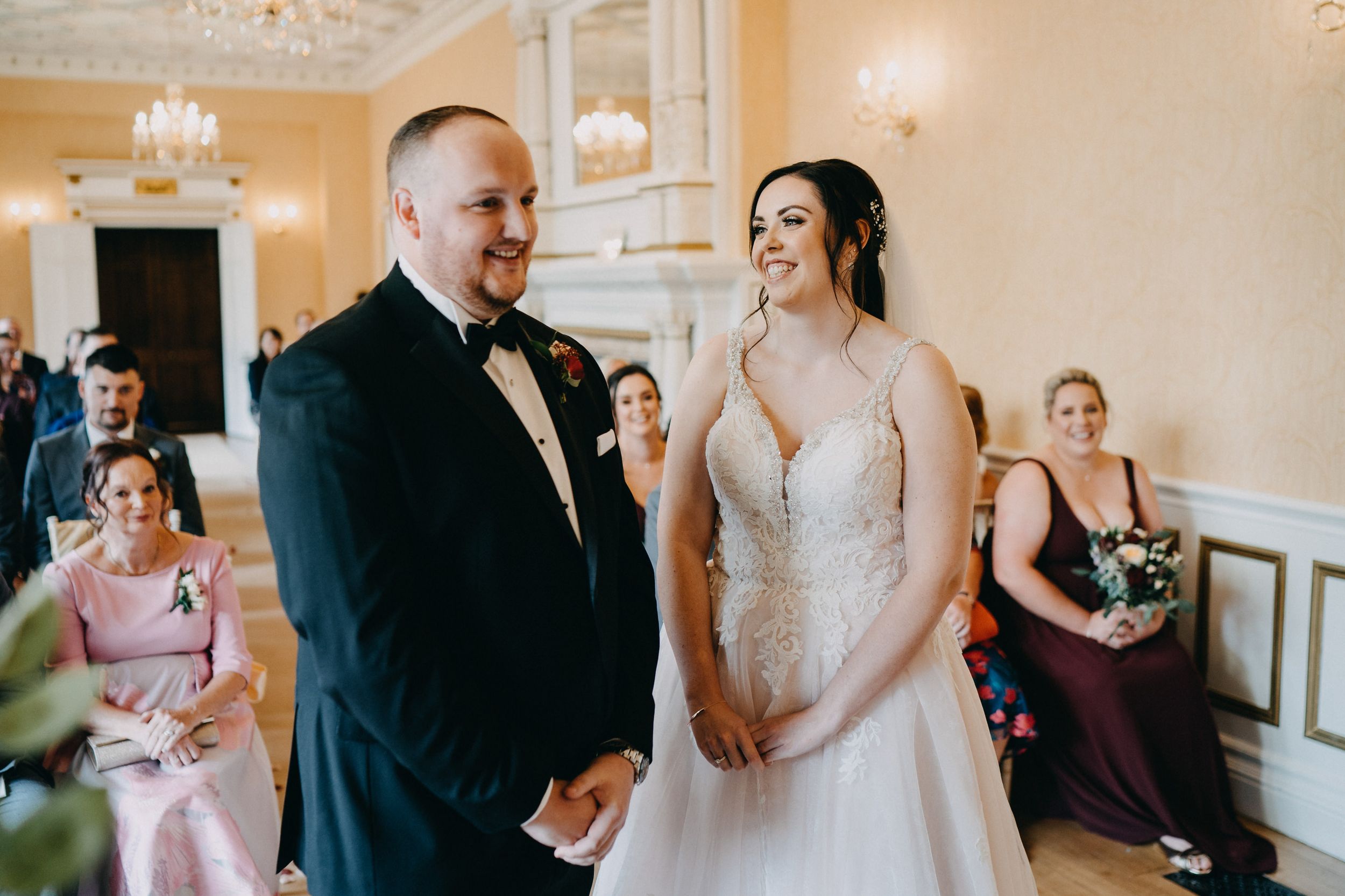 A joyful wedding couple sharing a laugh during their ceremony in an elegant ballroom with chandelier and white decor.
