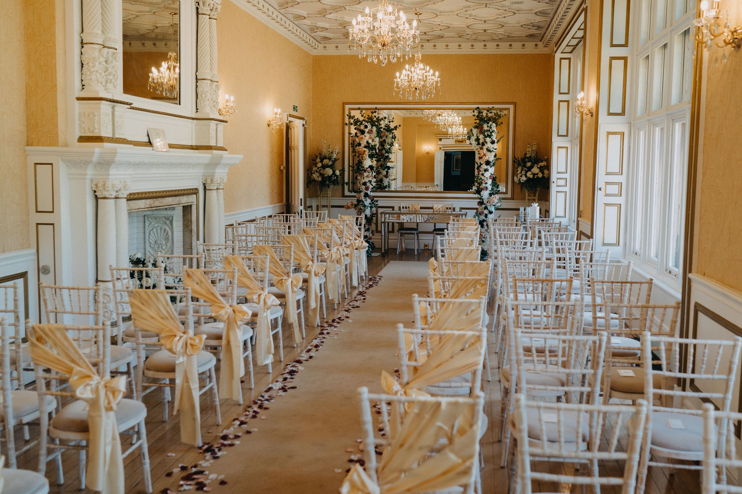 An elegant set up for a wedding ceremony at Holmewood Hall with rows of white chairs and crystal chandeliers hanging from ornate ceilings.