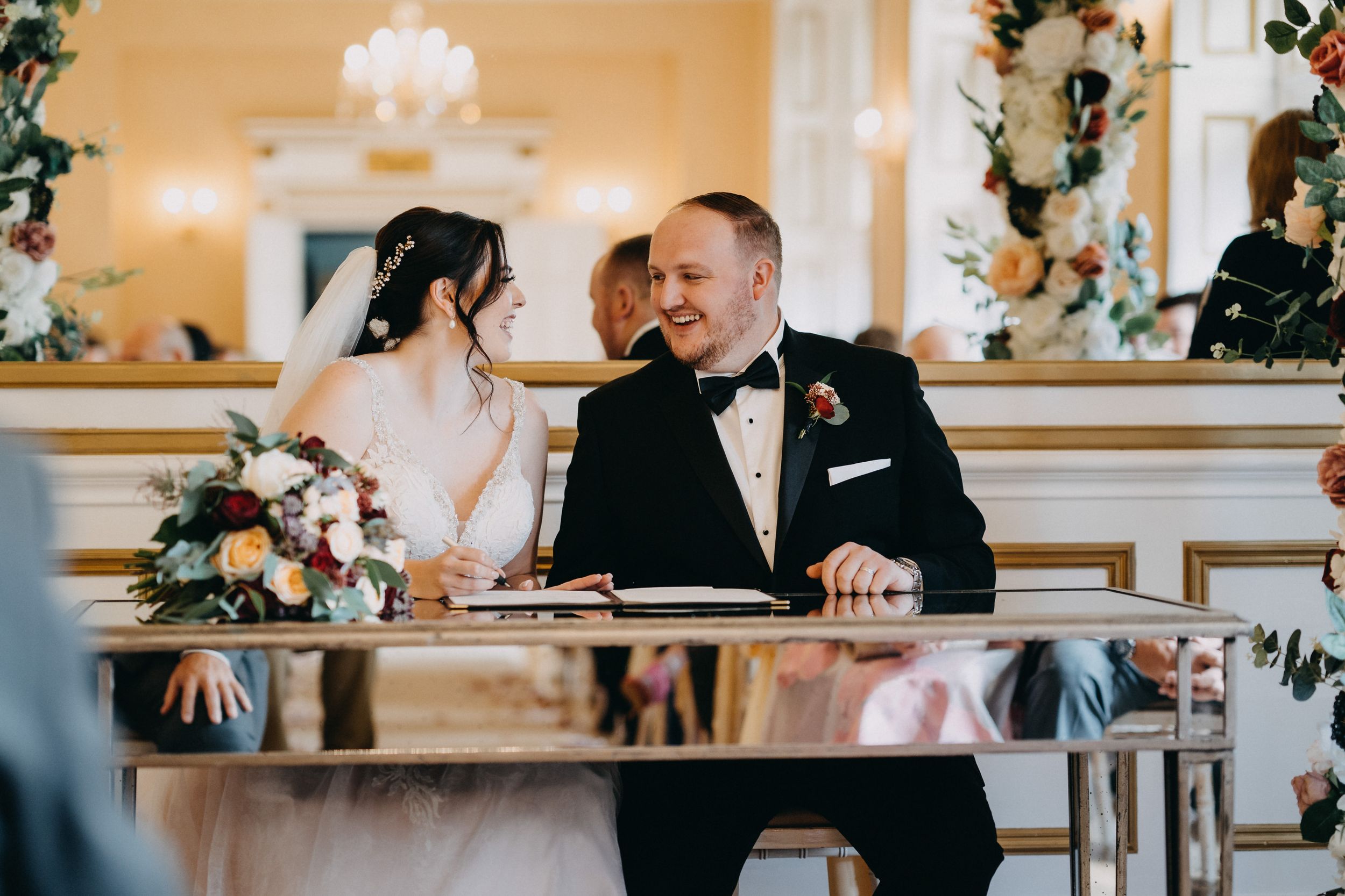 Elegant wedding scene at reception venue with couple seated at decorated table with floral arrangements and chandelier.