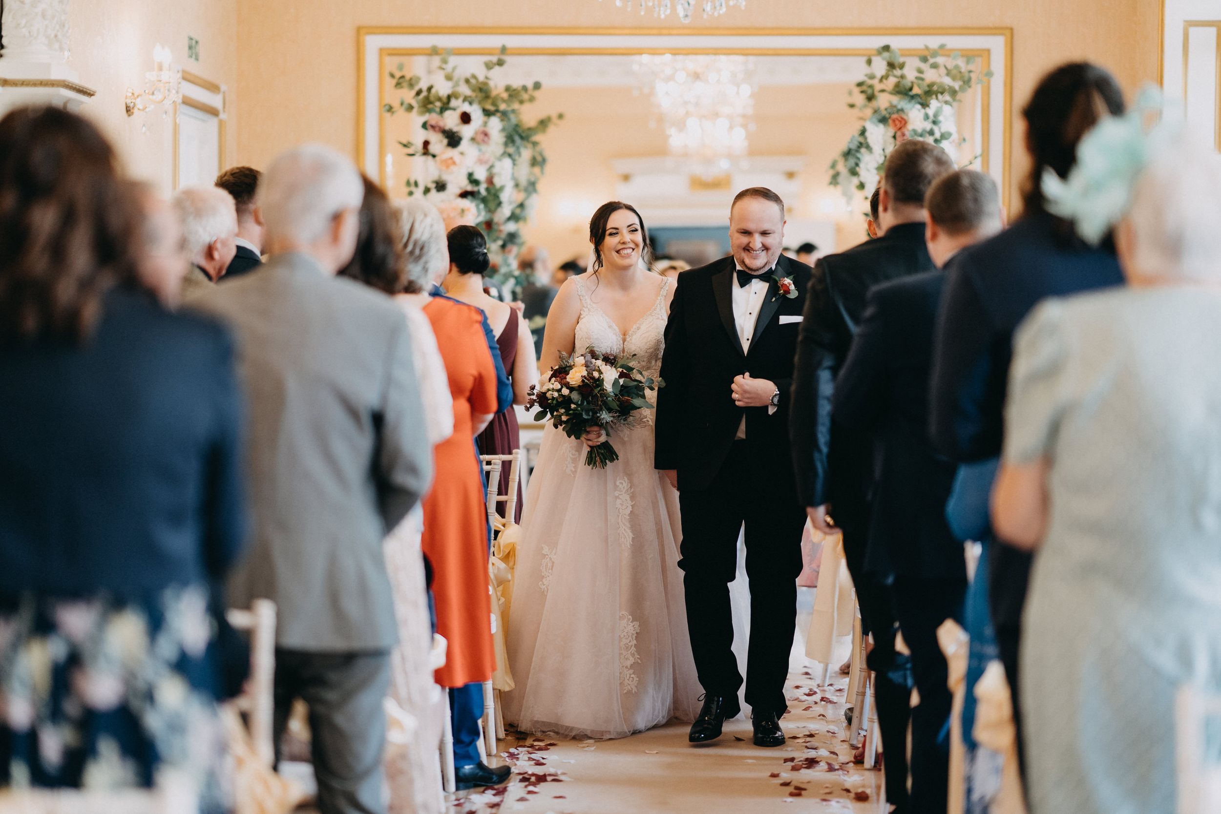 A couple walks down the aisle at an indoor wedding ceremony at Holmewood Hall surrounded by guests and floral decorations.