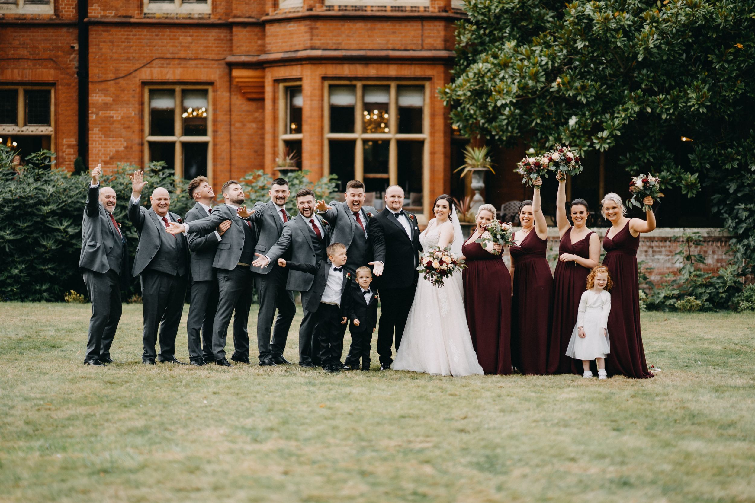 Wedding party poses on lawn in front of Holmewood Hall with groomsmen in gray suits and bridesmaids in burgundy dresses.