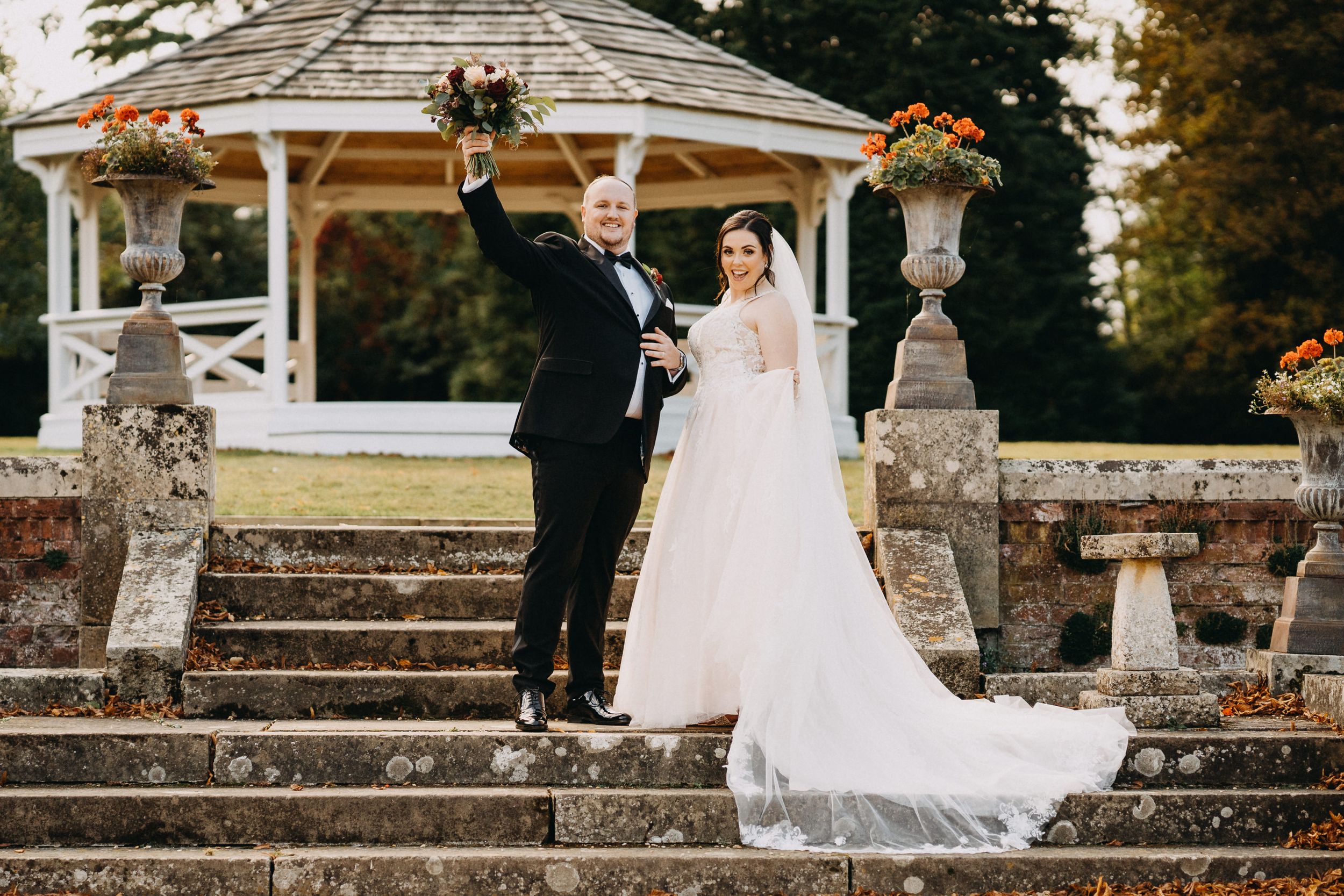 A romantic wedding couple stands on stone steps at Holmewood Hall in front of an ornate gazebo decorated with autumn flowers.