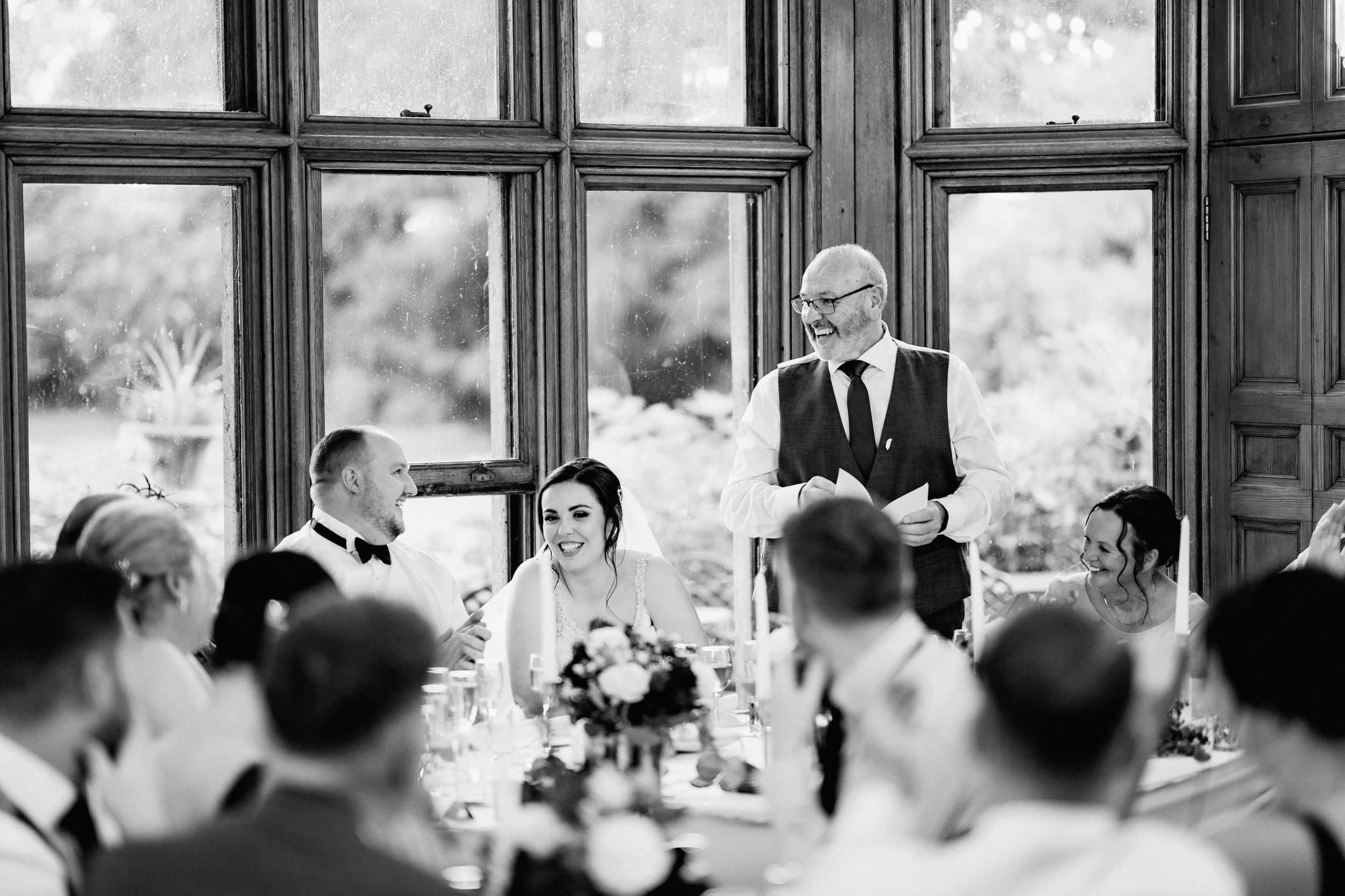 A black and white photograph of a wedding reception with people seated at tables listening to a formal toast.