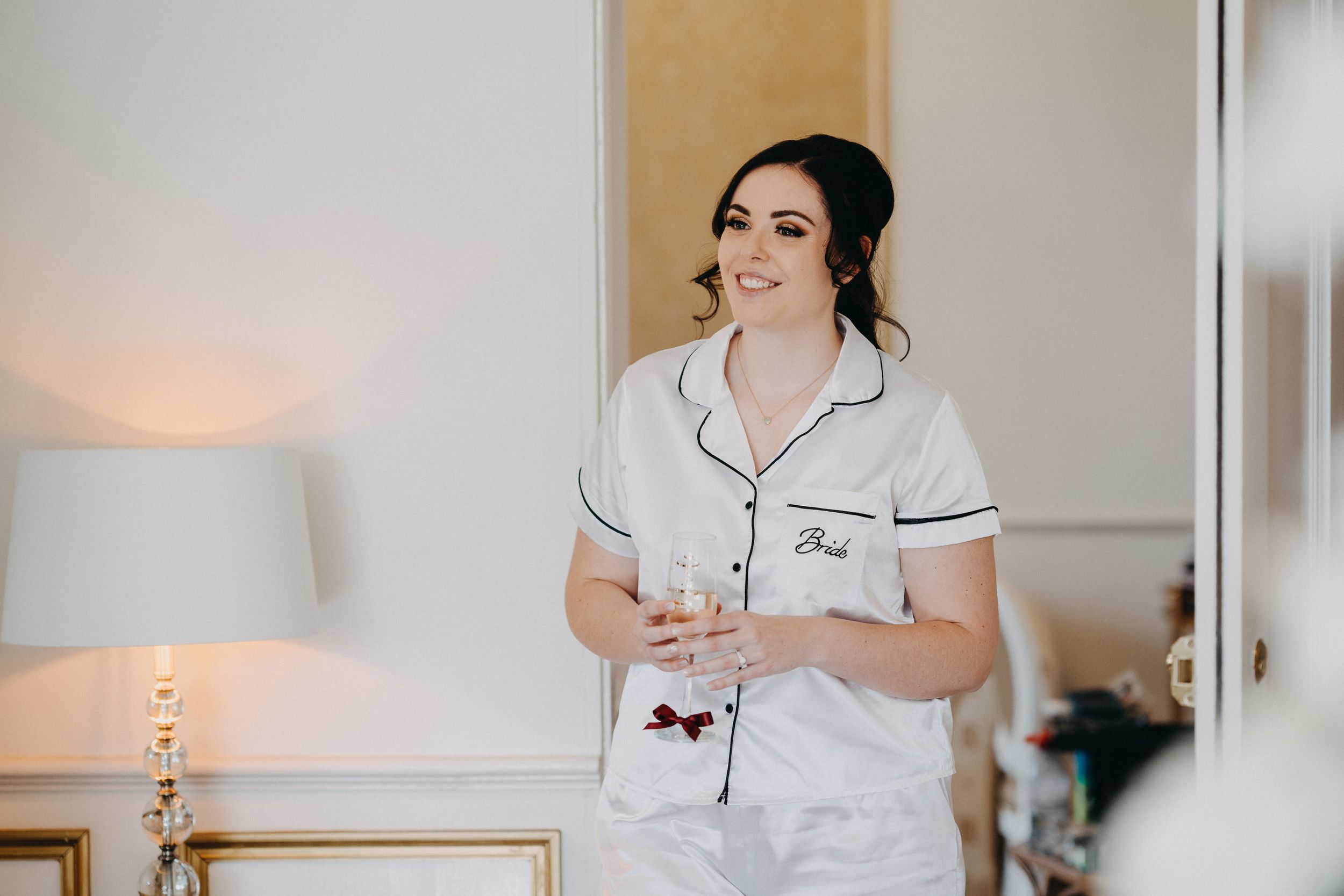 Someone wearing white silk pajamas with black piping holds a glass while smiling in a bedroom with warm lighting.