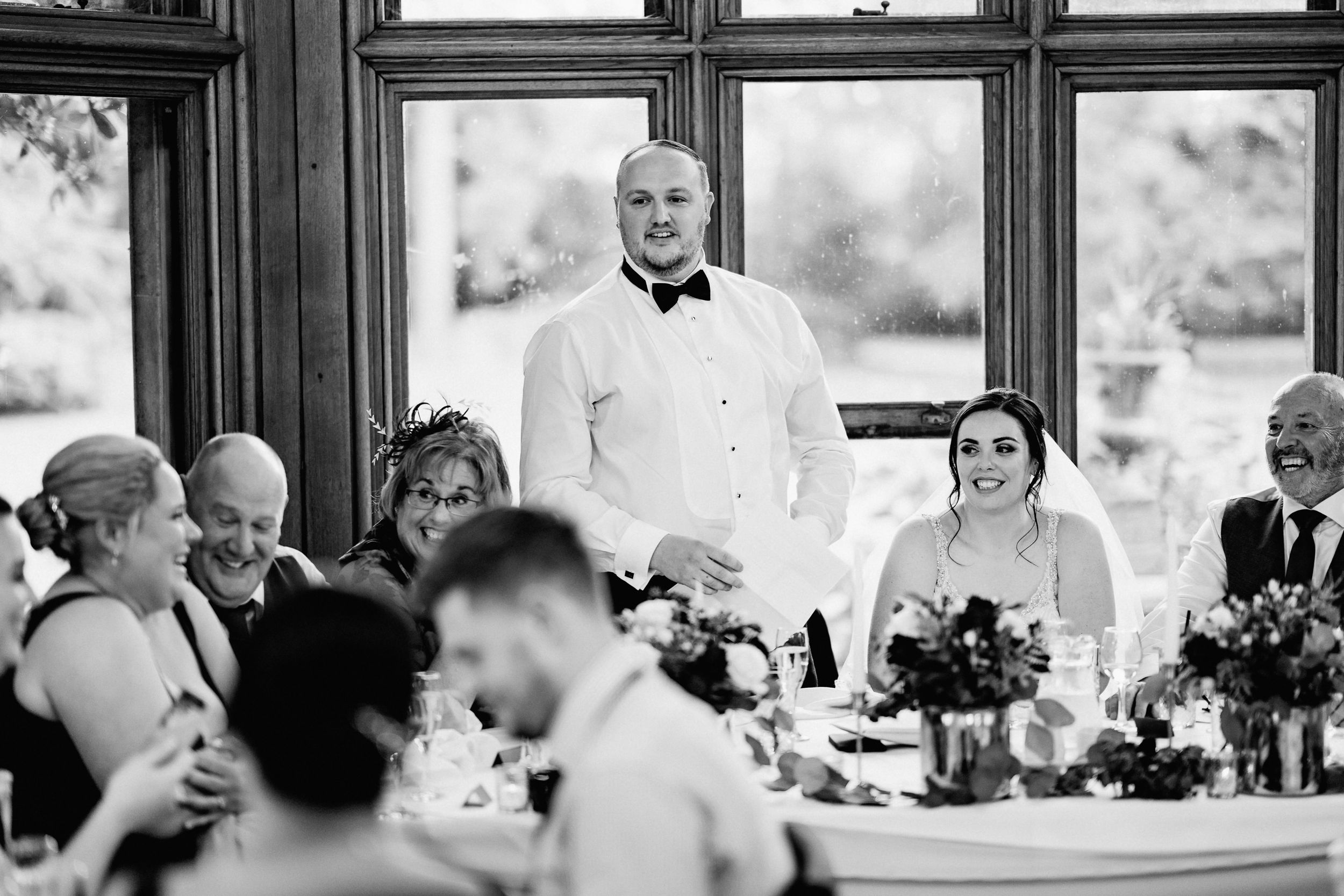 A black and white photograph of a wedding reception toast at an elegant venue with large windows.