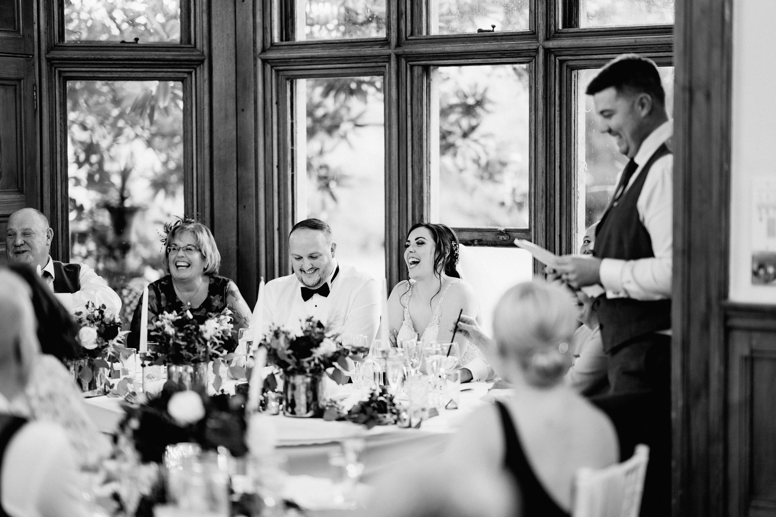 A black and white photo of a lively wedding reception with guests dining and laughing at an elegant indoor venue.
