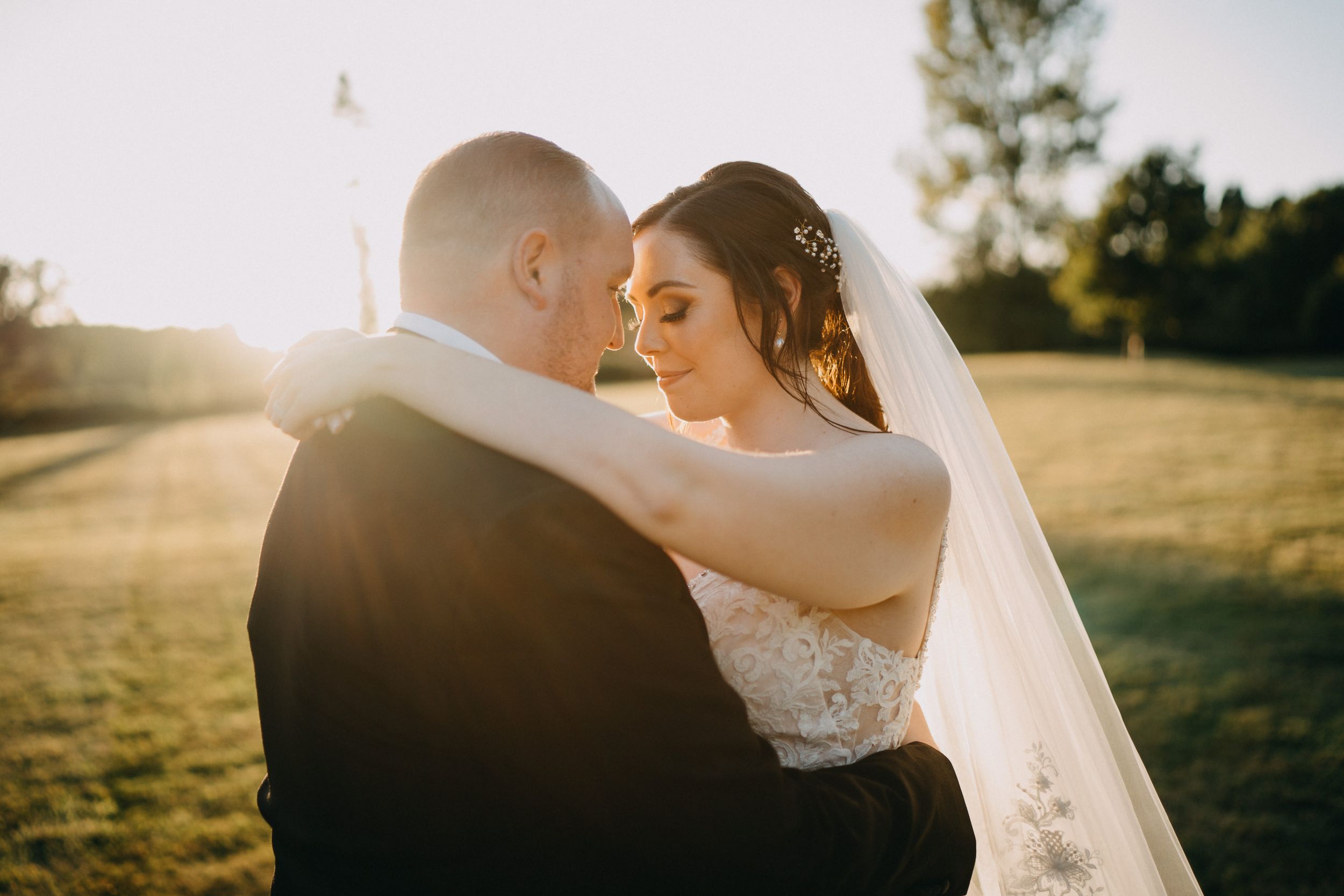 Romantic newlyweds embrace in golden sunlight on their wedding day in an outdoor field setting.