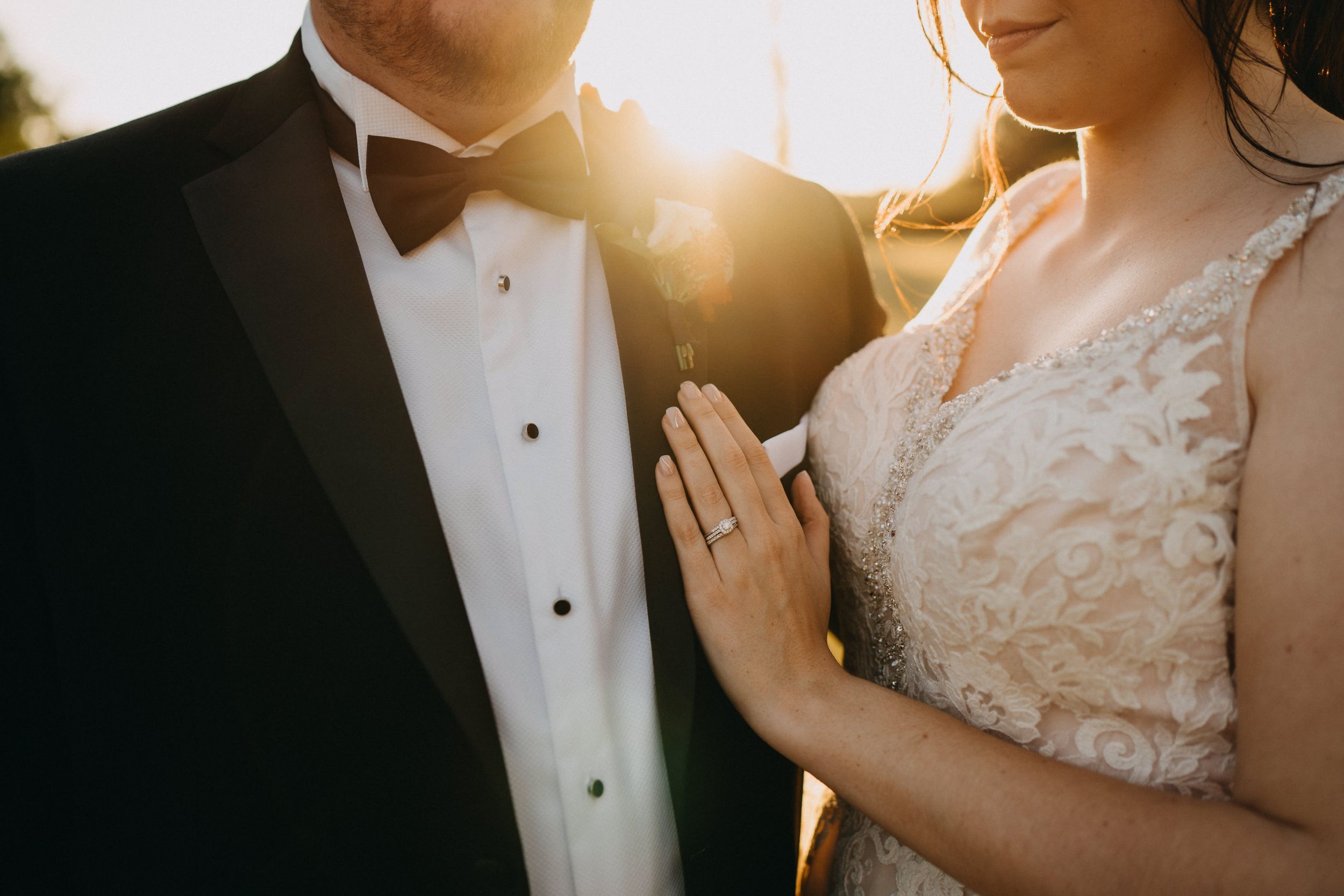 Newlyweds in formal attire stand together at golden hour with sunlight creating a romantic backlit glow.