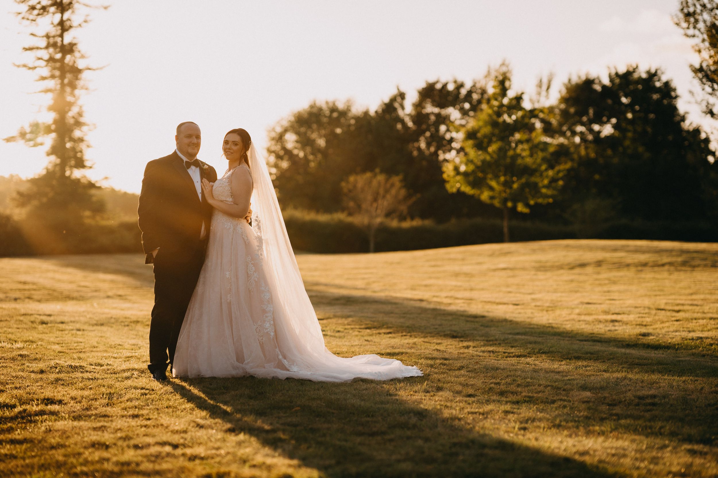 A couple at Holmewood Hall stands together on a grassy hill during golden hour sunset.