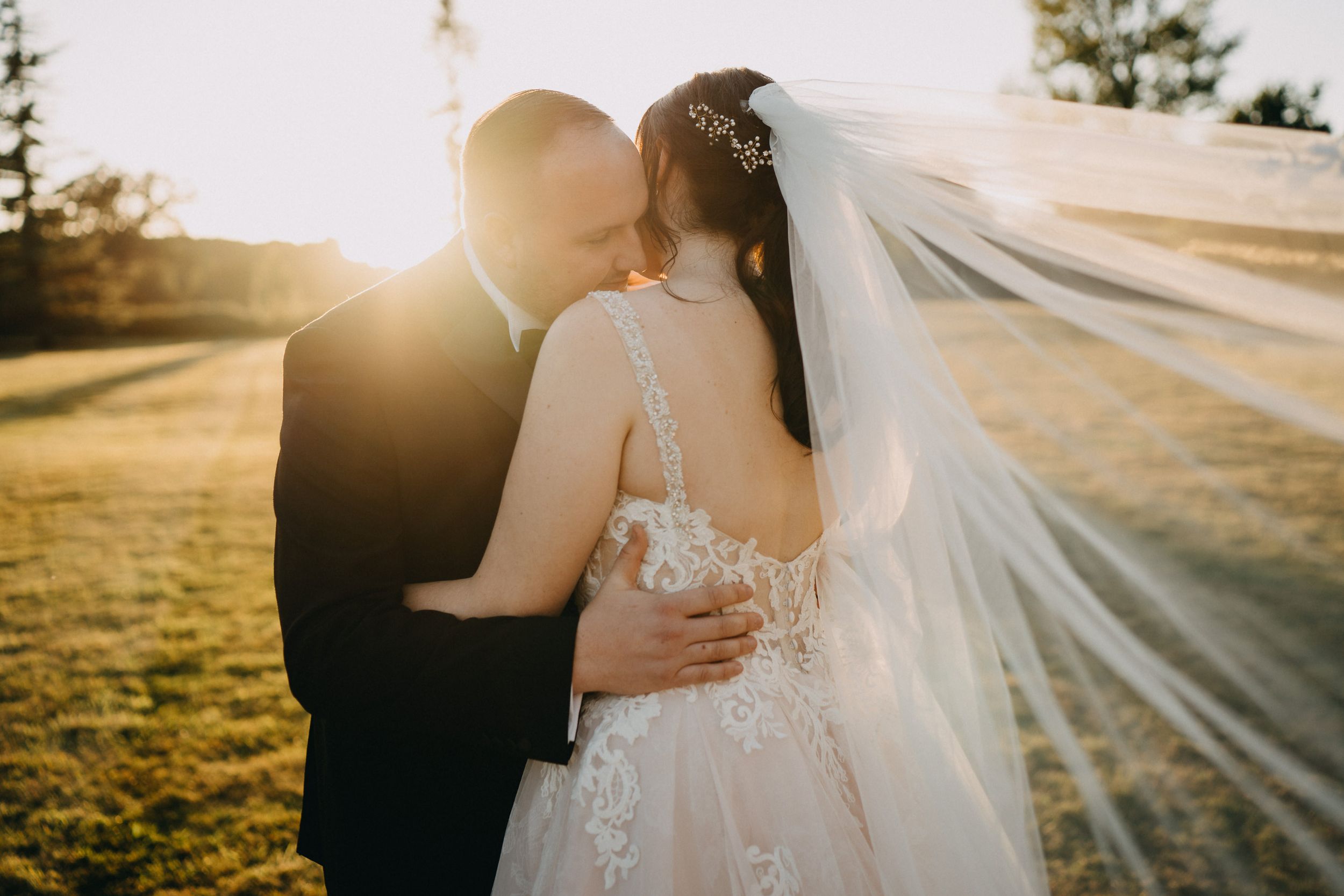 A romantic sunset wedding photo with a flowing veil and lace dress against golden sunlight in a grassy field.