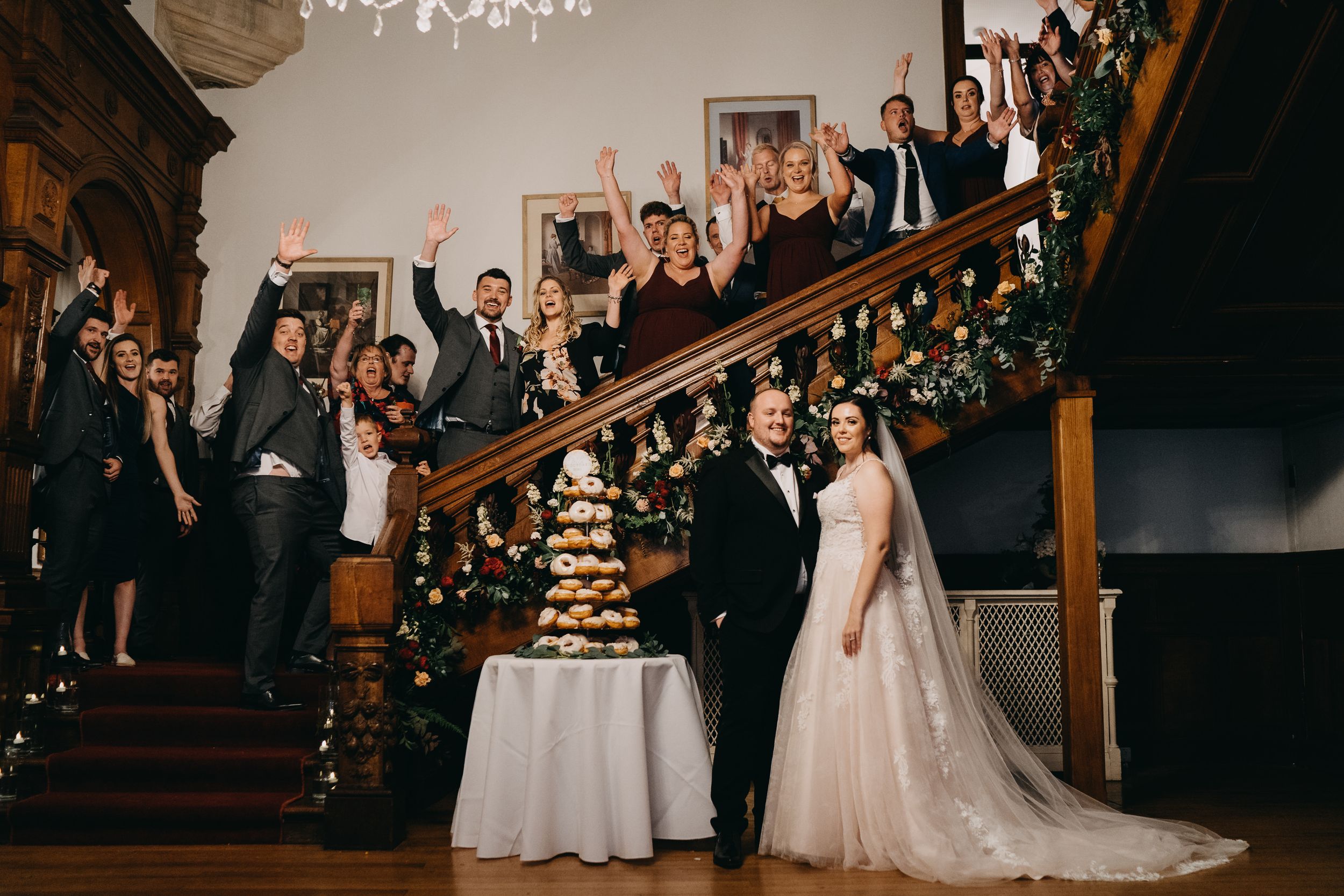 Wedding party celebrates on grand staircase with tiered cake at traditional elegant venue of Holmewood Hall, Peterborough.