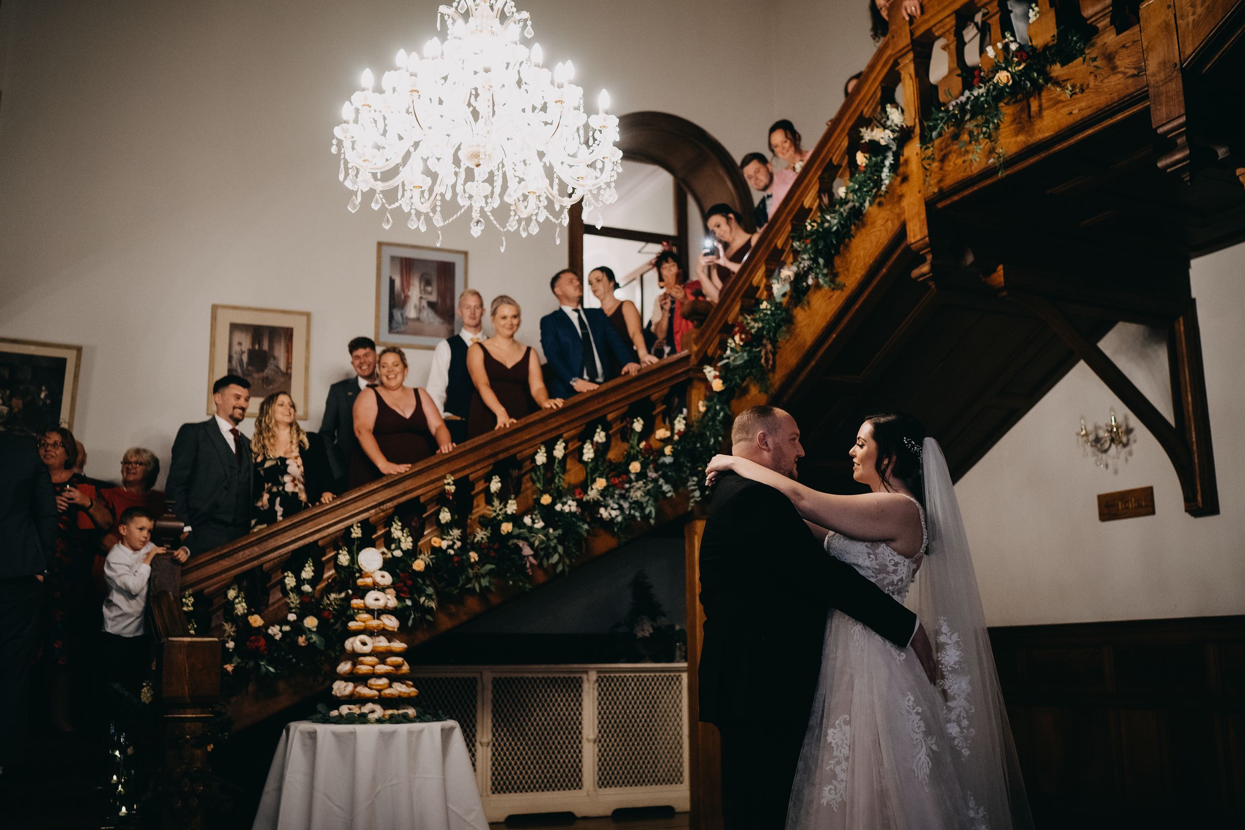 Wedding party elegantly arranged on grand wooden staircase with crystal chandelier at indoor venue.