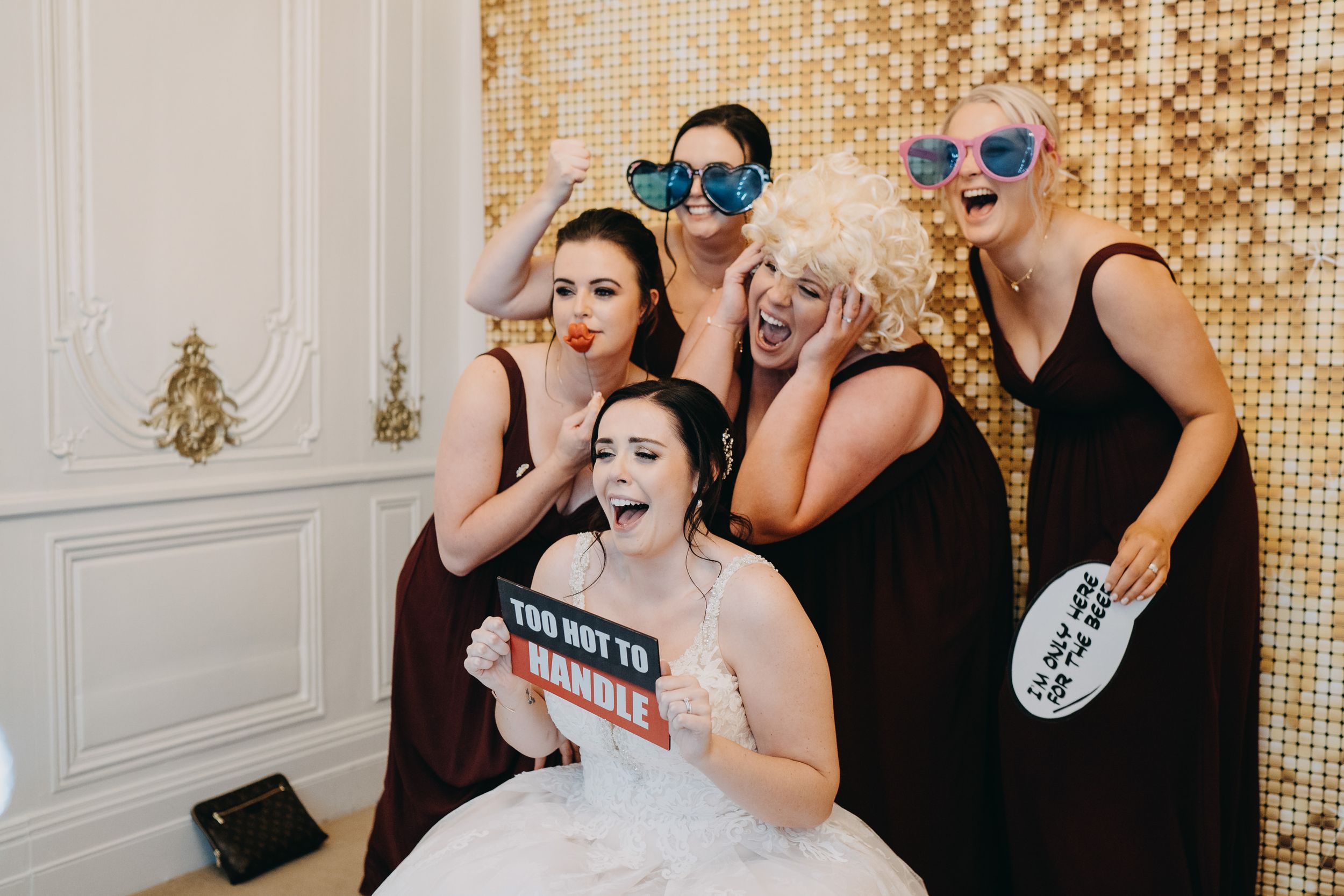 A group in formal attire poses playfully with props and sunglasses against a sparkly gold backdrop at a celebration.