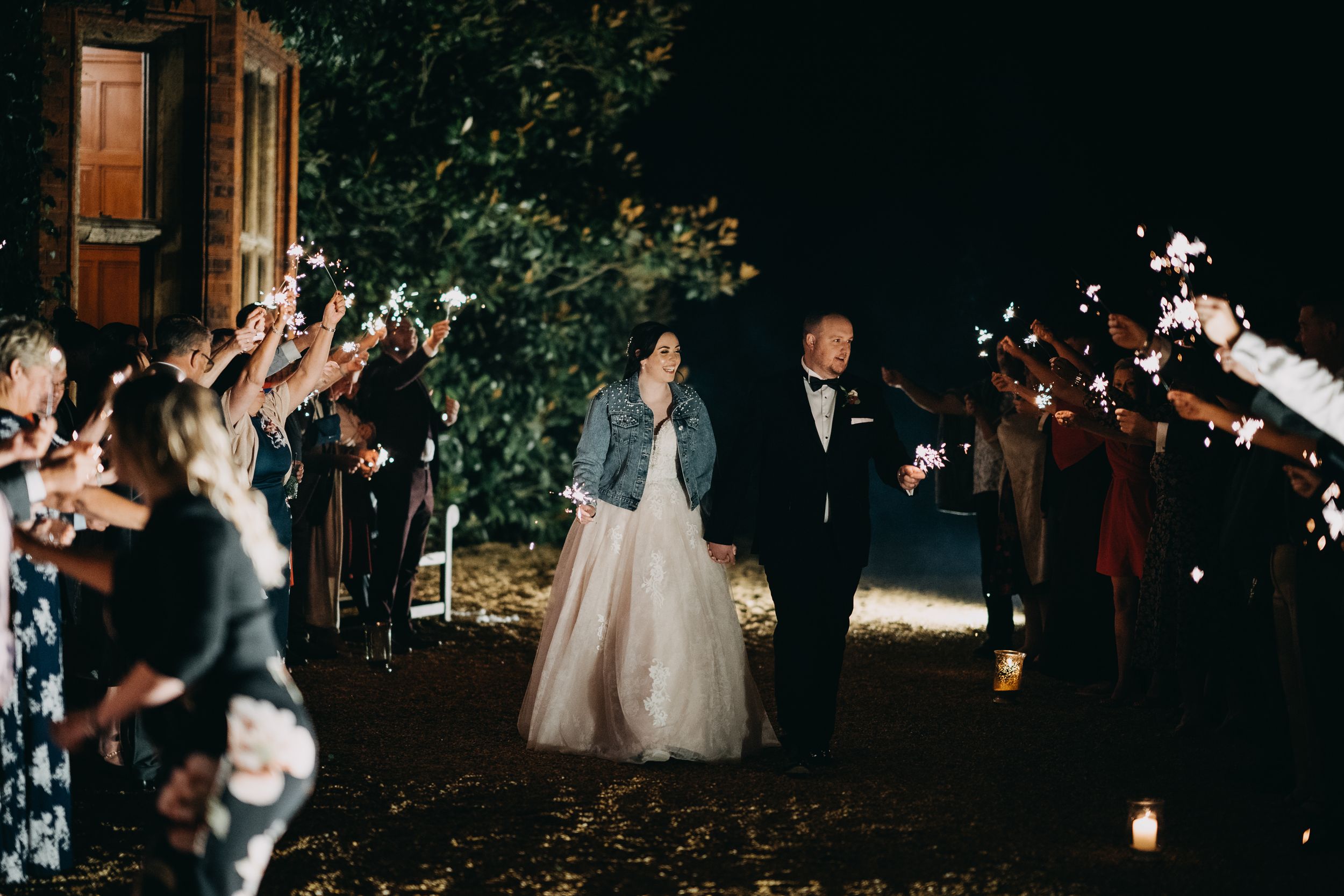 Wedding couple walks through sparklers at Holmewood Hall during nighttime departure as guests celebrate at outdoor venue.